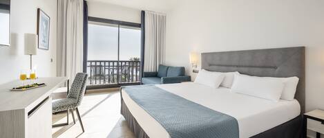 A modern hotel room with a large bed, a desk with a chair, a balcony with a view, and a dining area with a table and chairs.