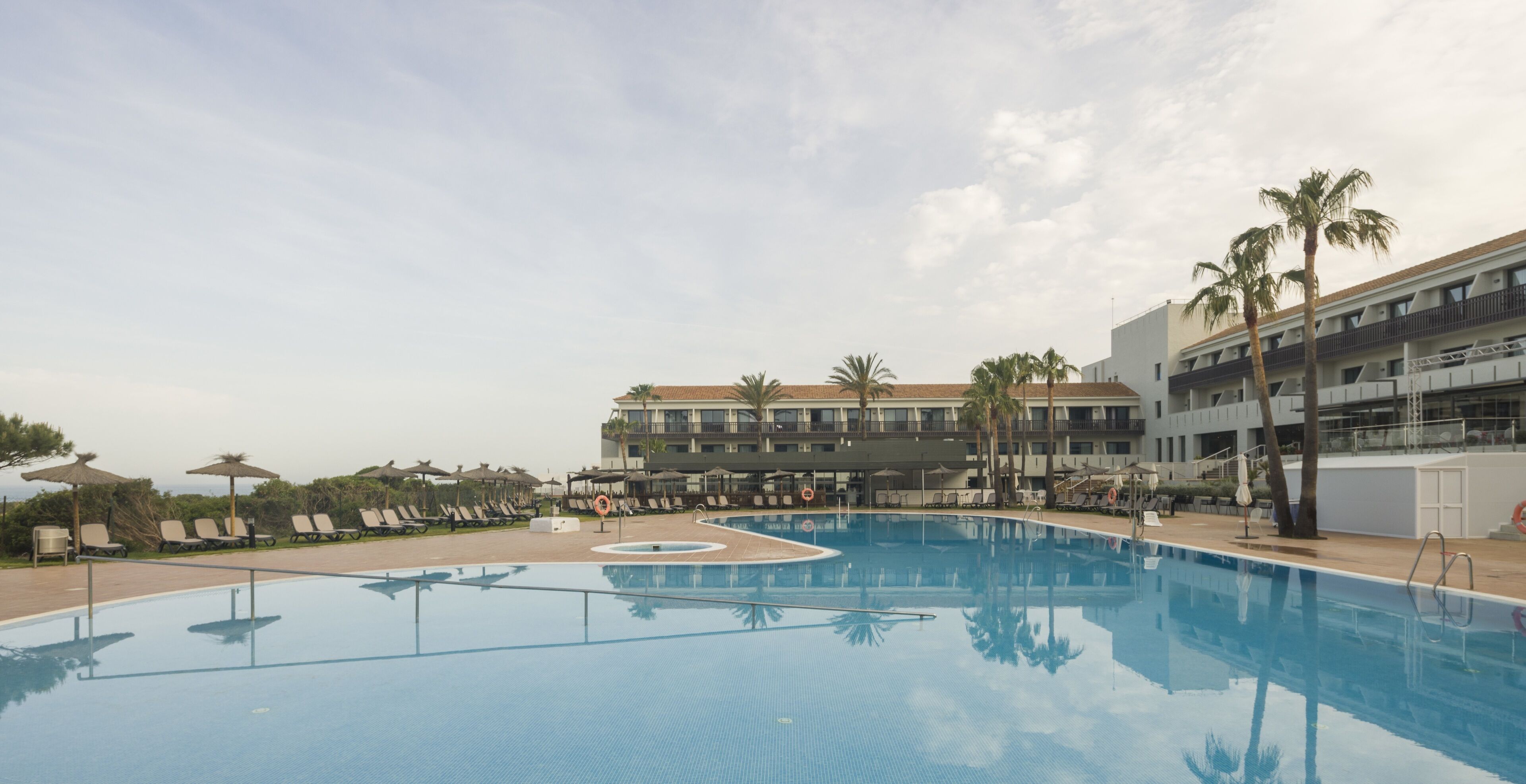 A swimming pool with lounge chairs and umbrellas, surrounded by palm trees and a building with balconies.