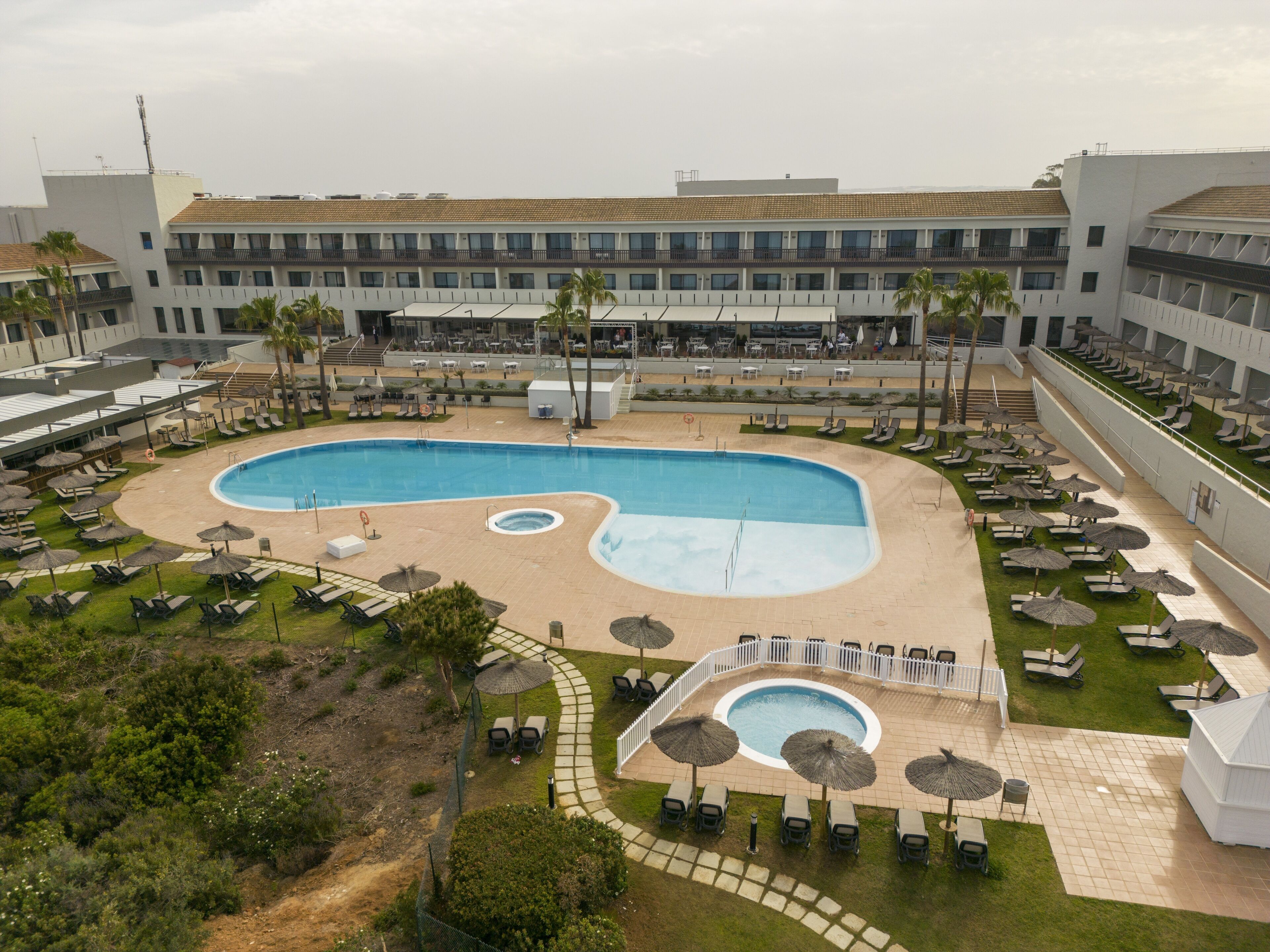 Ein großer Pool mit ungewöhnlicher Form, umgeben von Liegestühlen und Sonnenschirmen, im Hintergrund ein mehrstöckiges Gebäude.