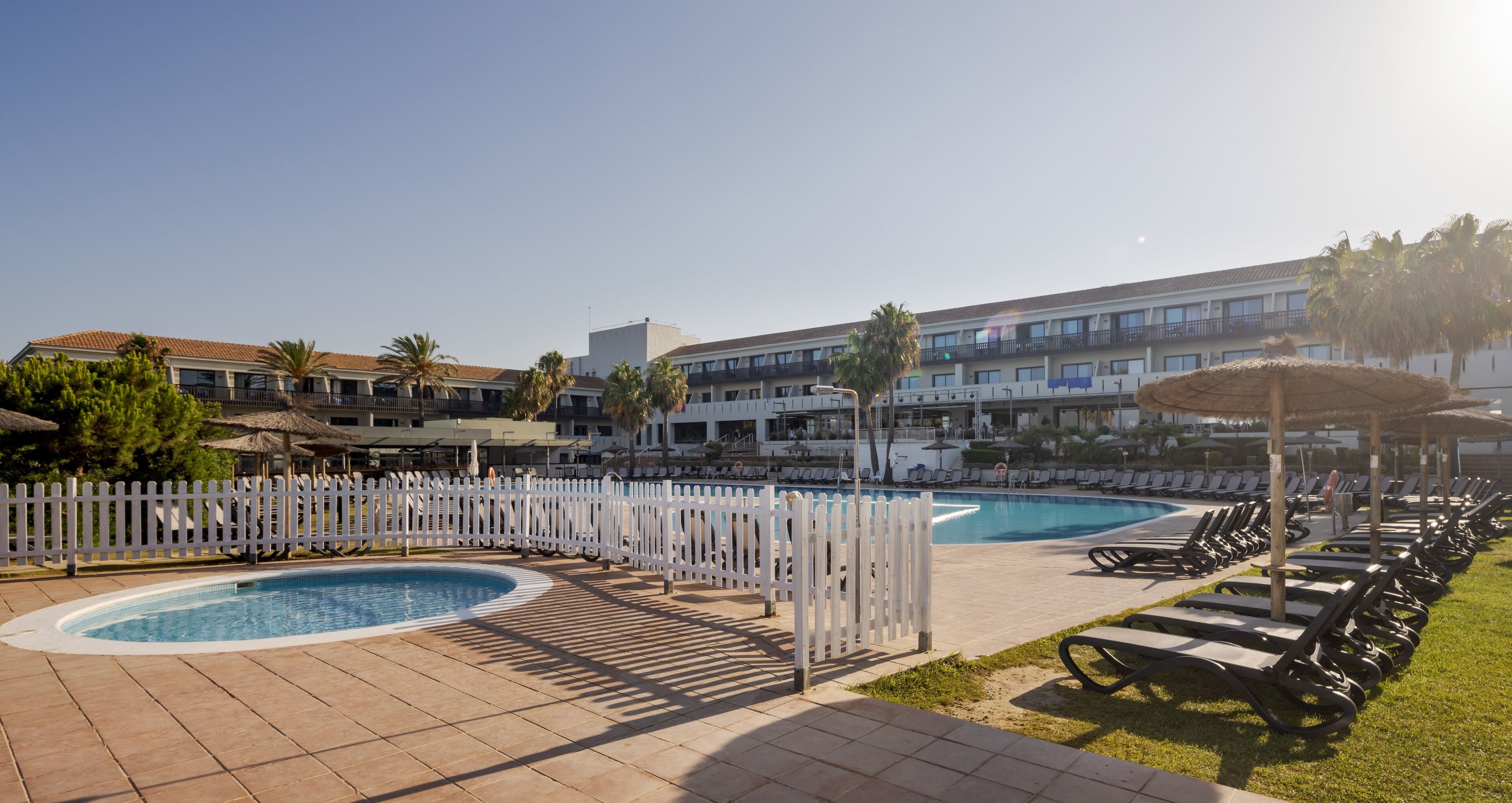 Zwembadgedeelte van een resort, met een witte omheining, ligbedden en een gebouw op de achtergrond.