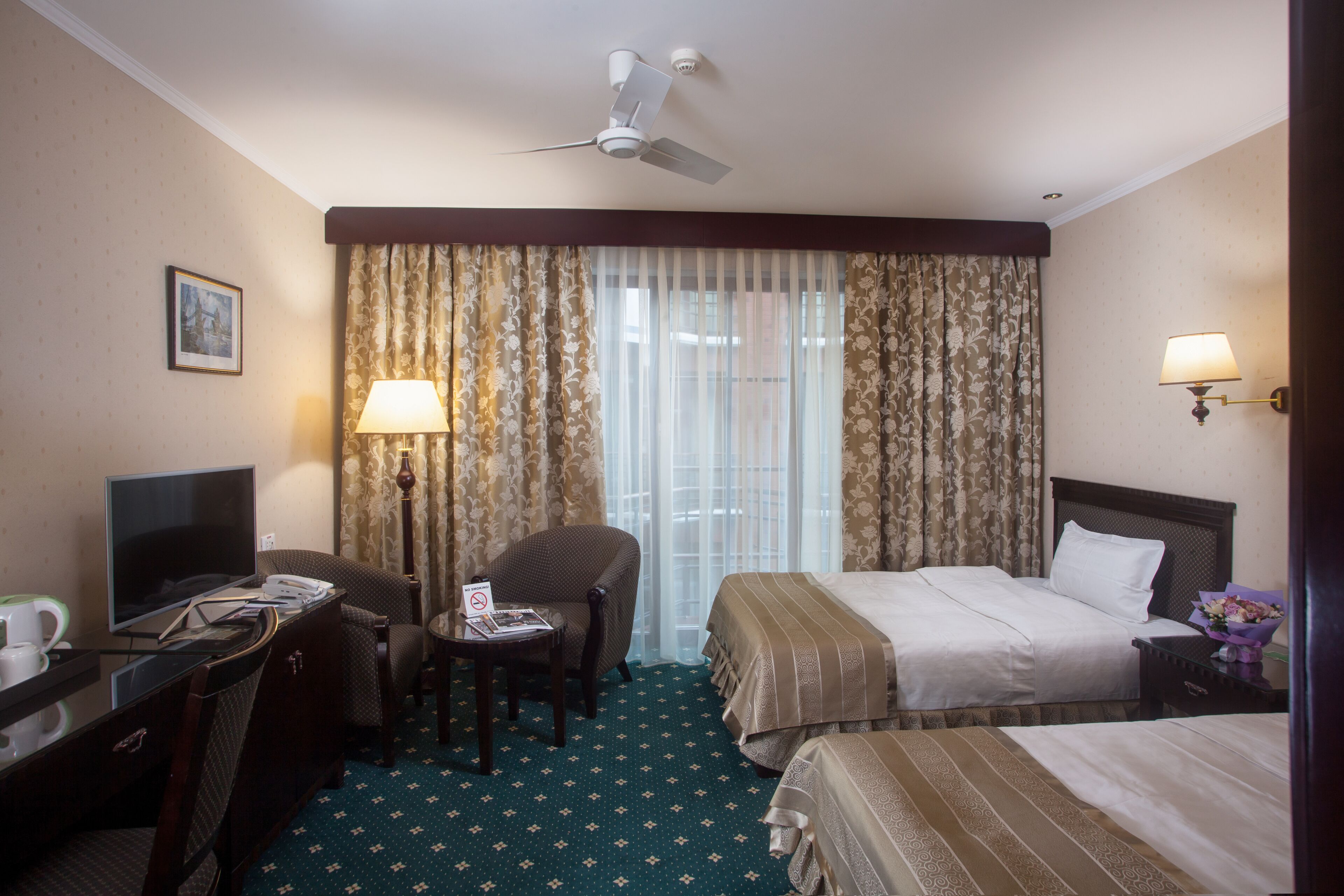 Een hotelkamer met een bed, een bureau met een computer, een stoel, een televisie en een plafondventilator.