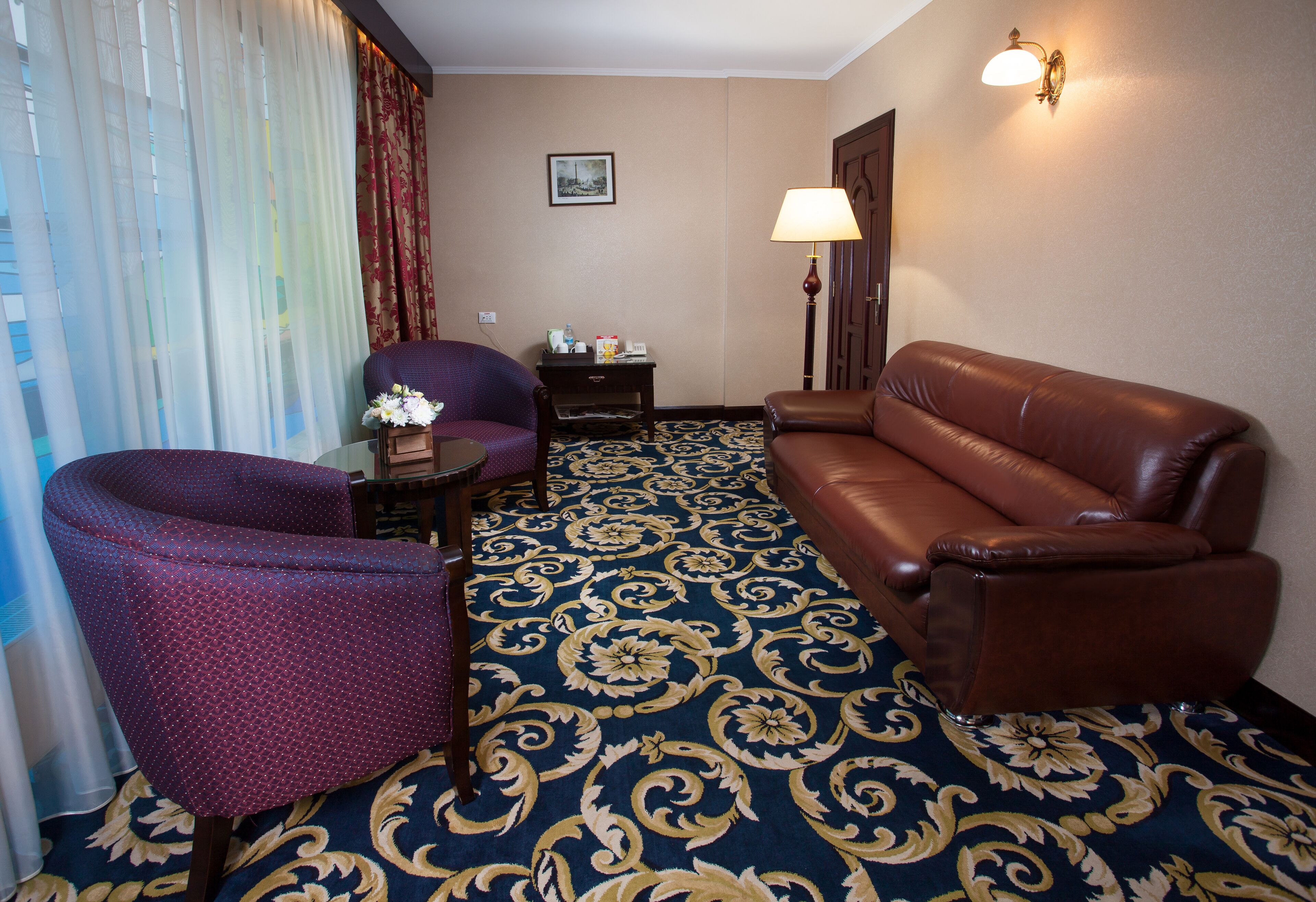 A hotel room with a patterned carpet, a brown leather sofa, a purple armchair, a small table with a vase of flowers, and a lamp.