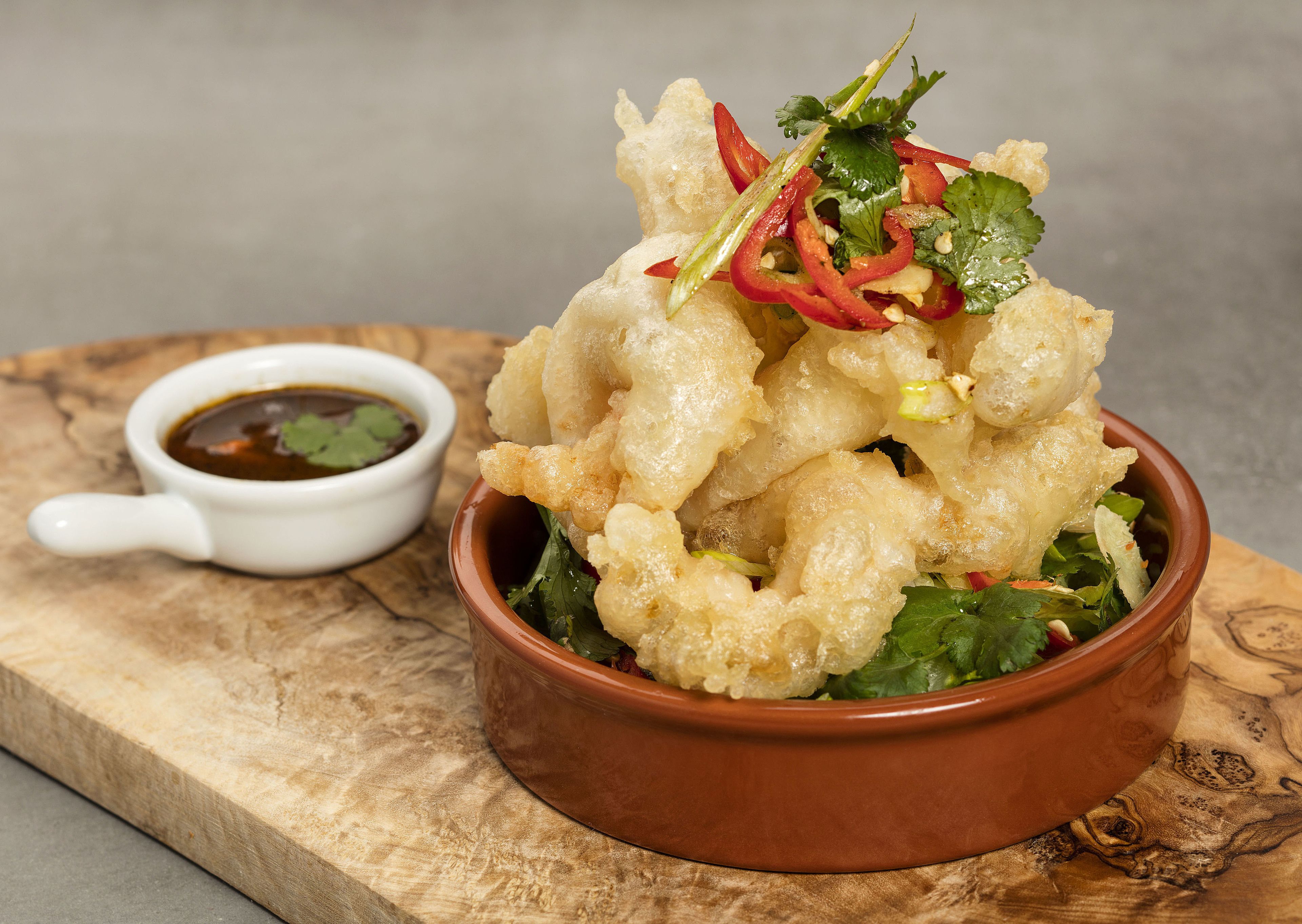 A bowl of crispy fried food garnished with vegetables, accompanied by a small bowl of dipping sauce on a wooden board.