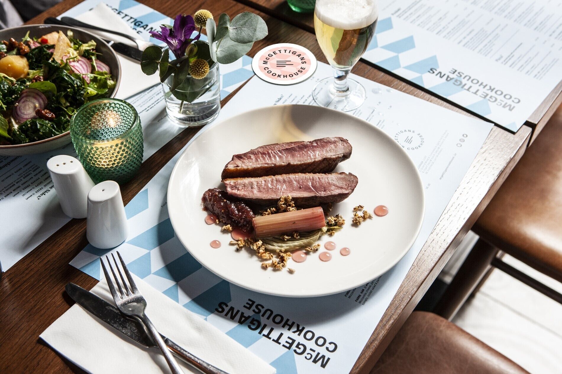 Une assiette de magret de canard avec sa sauce et des légumes, un verre de bière et un menu sur une table en bois.