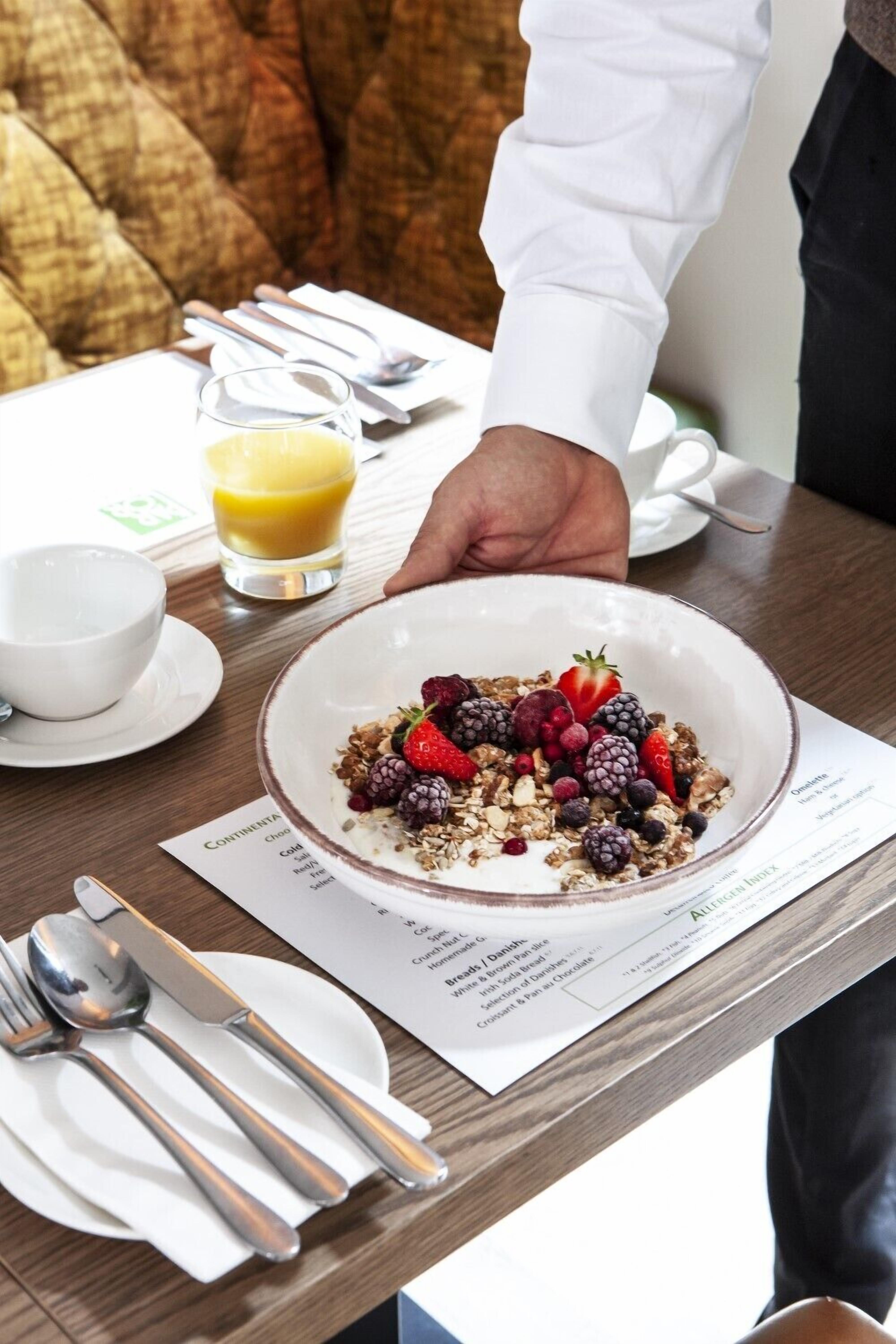 A breakfast setting with a bowl of cereal topped with berries, a glass of orange juice, and a menu on the table.