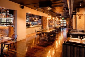 L’intérieur d’un restaurant présente un long bar en bois, plusieurs tables et un mur en briques.