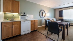 Cocina compacta con muebles de madera, lavavajillas blanco y microondas. Hay una zona de comedor con mesa y sillas, y una sala de estar con sofá y un espejo redondo en la pared.