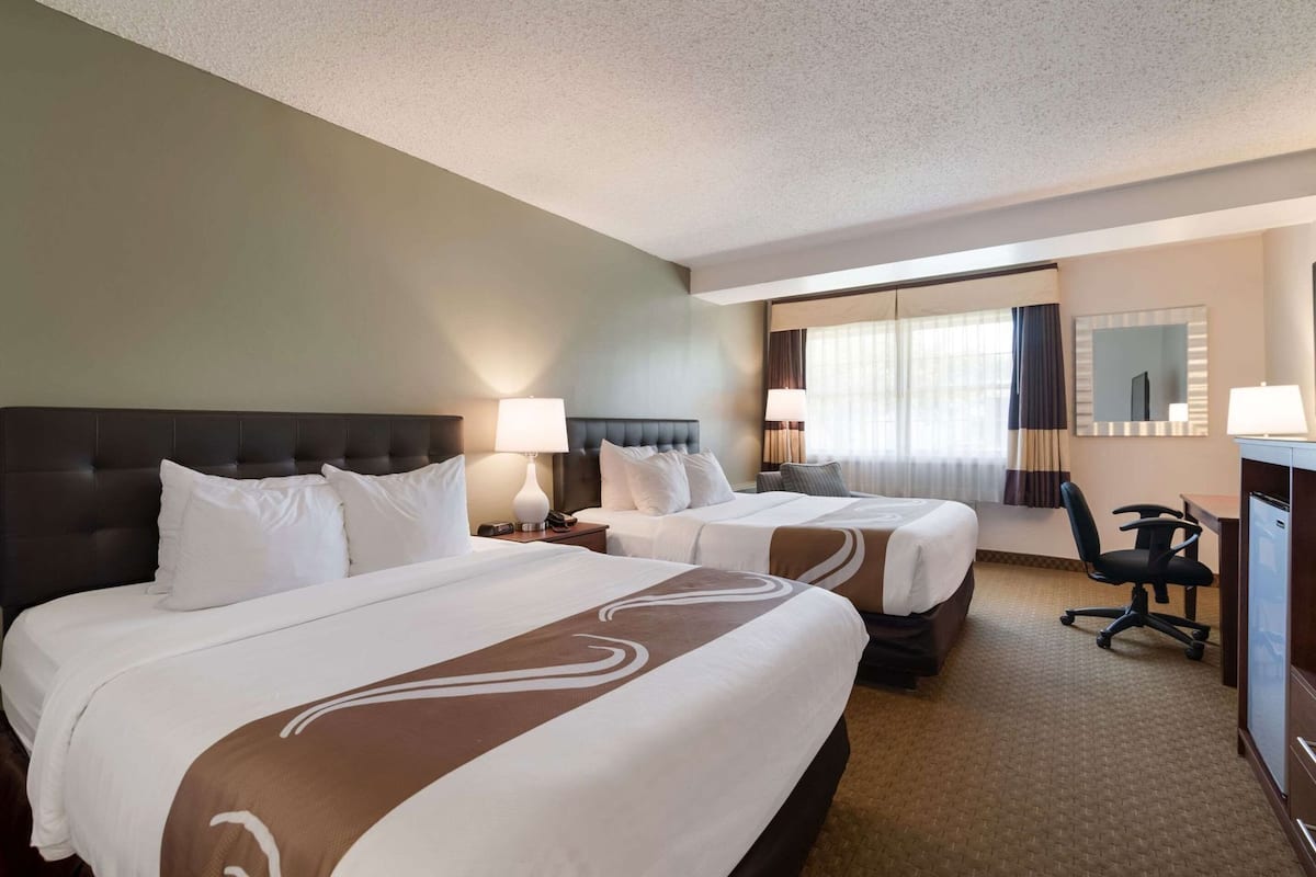 Quarto de hotel com duas camas, uma escrivaninha, uma cadeira e um espelho.