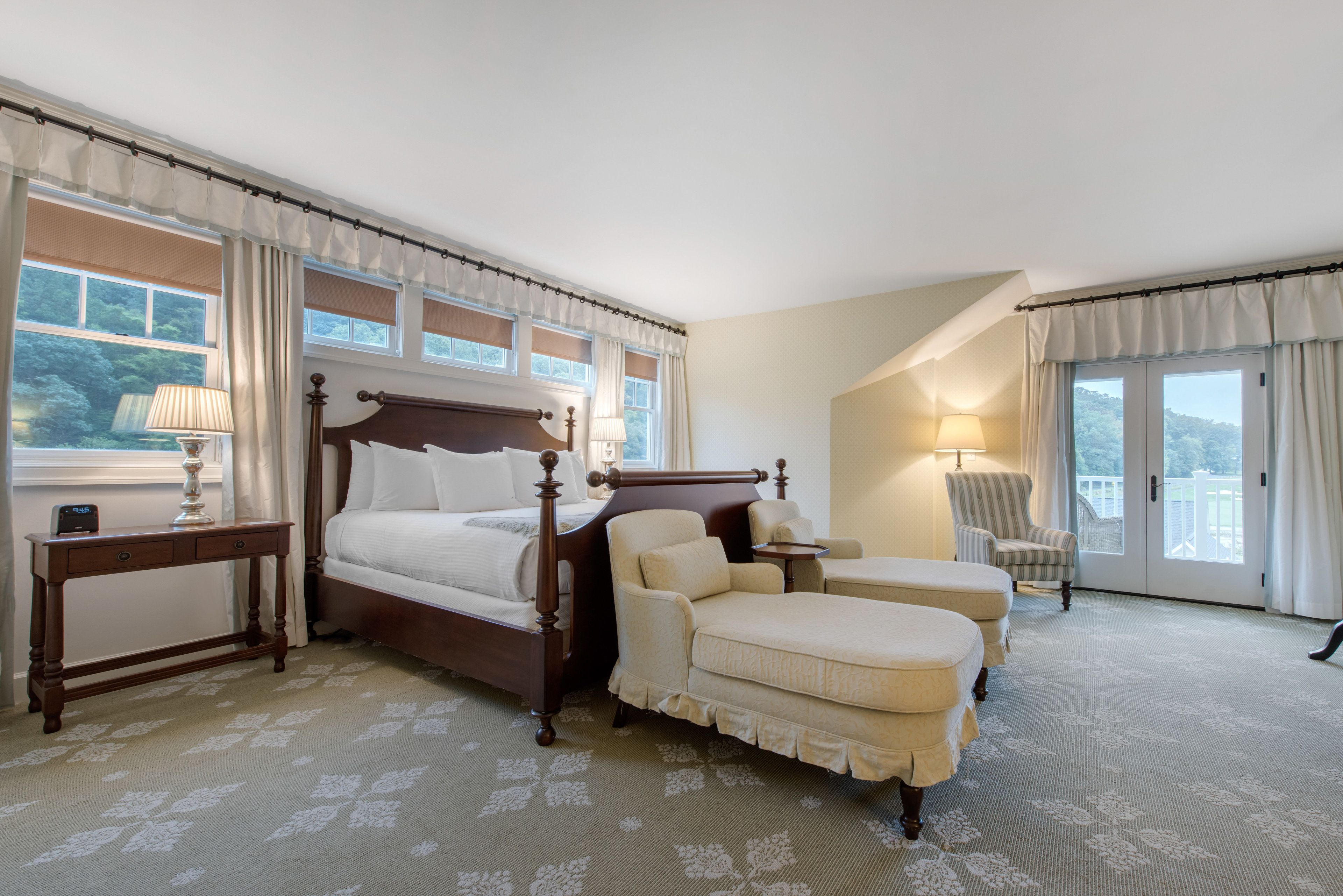 Quarto de hotel com cama de dossel, área de estar com poltrona e apoio para os pés, mesa pequena com abajur e vista para o exterior por grandes janelas.