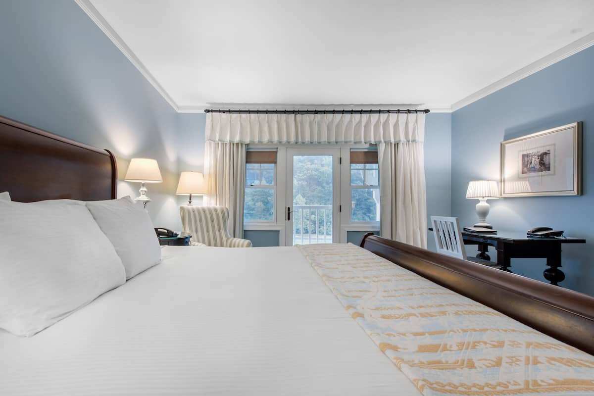 Ein Hotelzimmer mit einem großen Bett, einem Schreibtisch mit Lampe, einem Sessel und Blick ins Freie durch eine Tür.