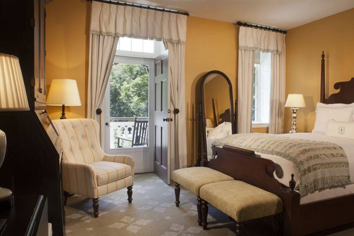 Ein Schlafzimmer mit einem großen Bett, zwei Nachttischlampen, einem Sessel, einer Bank, einem Spiegel und einer Tür zum Balkon.