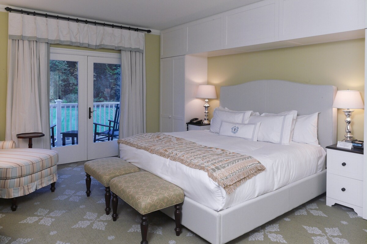 A bedroom with a large bed, a bench, a nightstand, and a view through a door to an outdoor area.