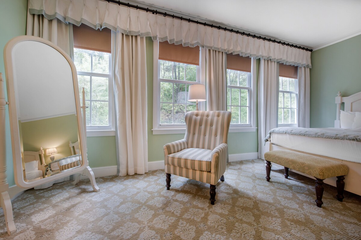 A bedroom with a large mirror, a striped armchair, a bench, and a bed.