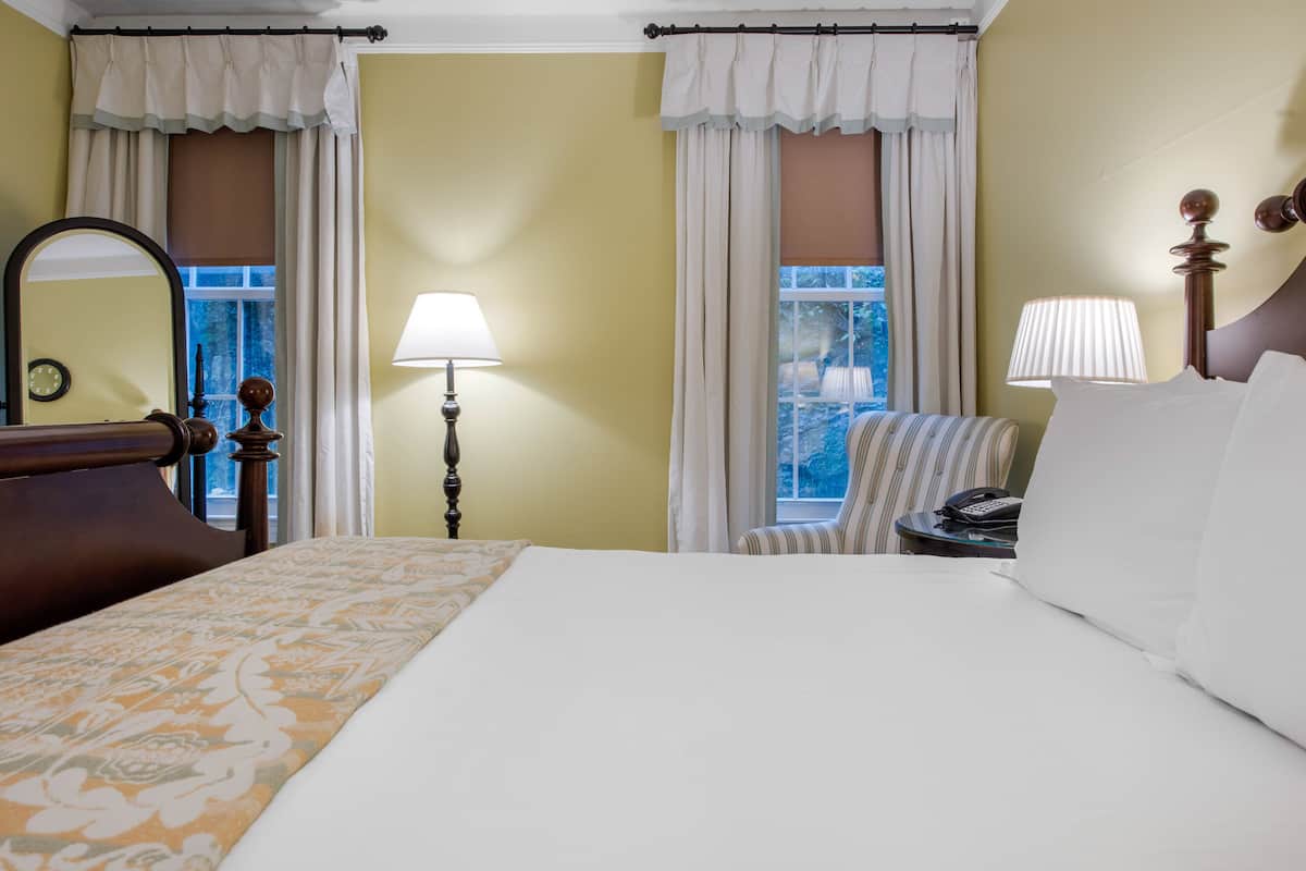 Ein Hotelzimmer mit einem großen Bett, zwei Nachttischlampen, einem Sessel und einem Fenster mit Vorhängen.