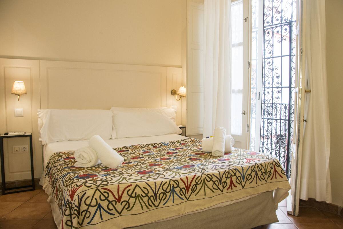 Habitació d'hotel amb un llit, dues làmpades de nit, una taula de nit i una finestra amb reixatge metàl·lic.