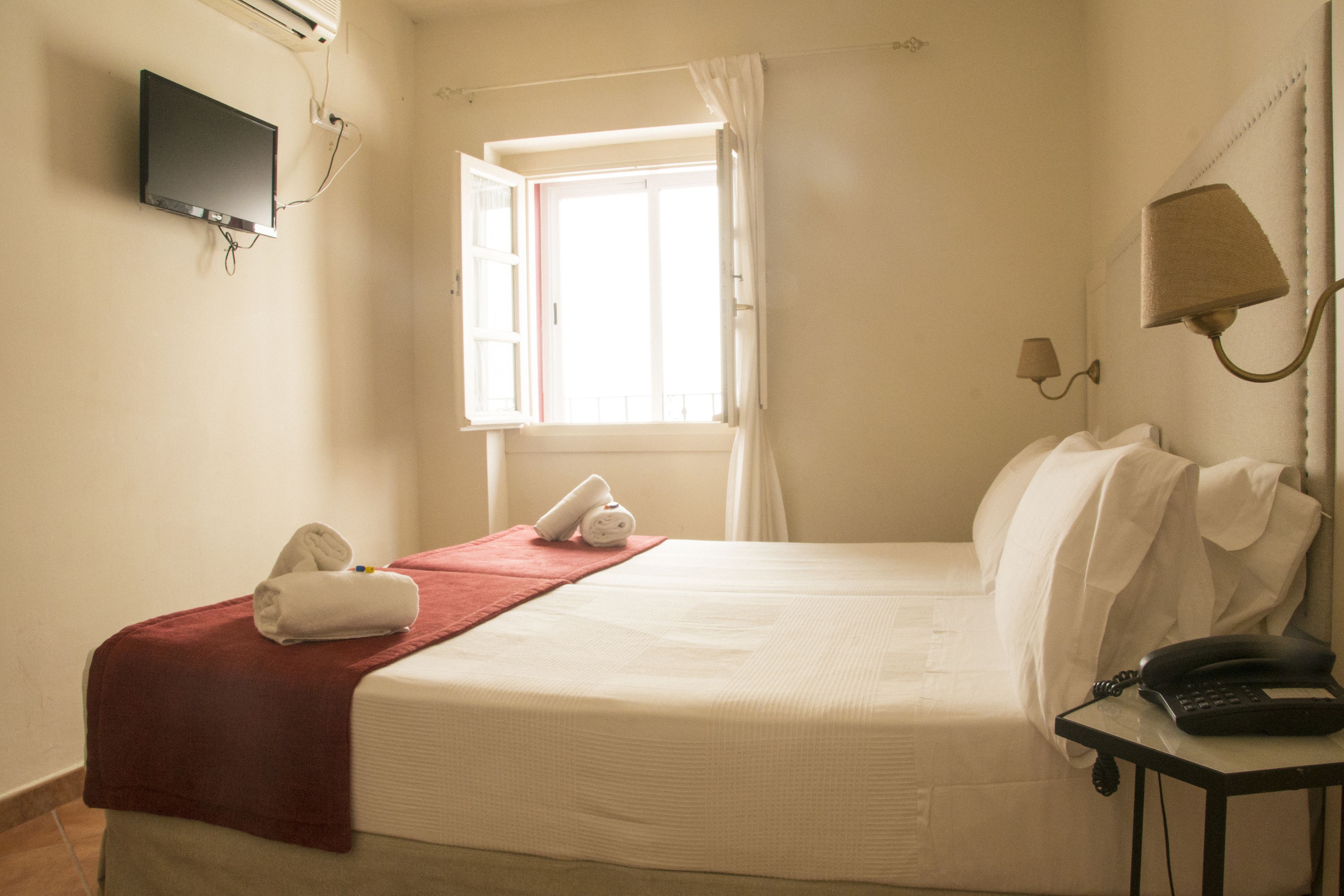 A hotel room with a bed, a TV mounted on the wall, a window with curtains, a bedside lamp, and a telephone on a small table.