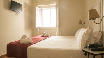 A hotel room with a bed, a TV mounted on the wall, a window with curtains, a bedside lamp, and a telephone on a small table.