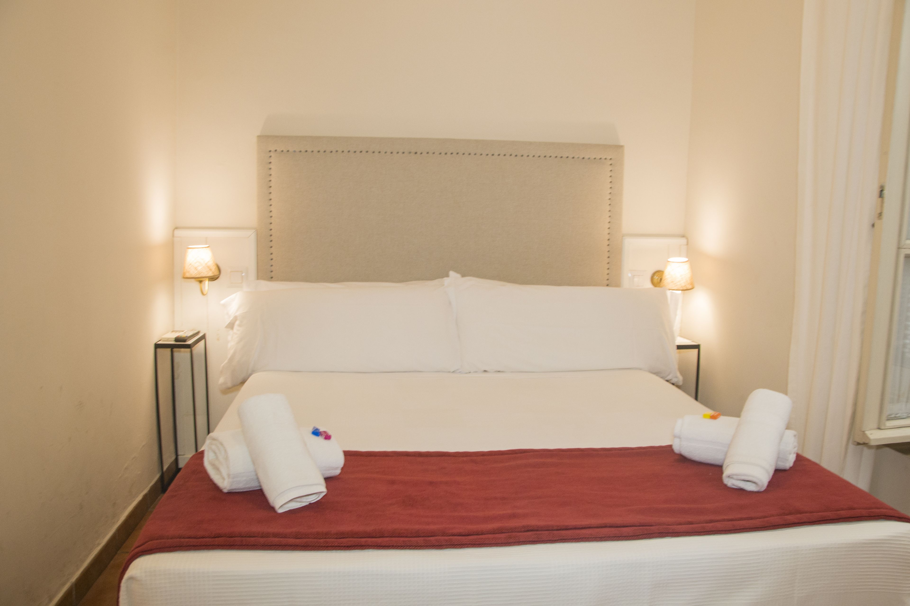 Una habitación de hotel con una cama, dos lámparas de noche, una manta roja y dos toallas enrolladas sobre la cama.