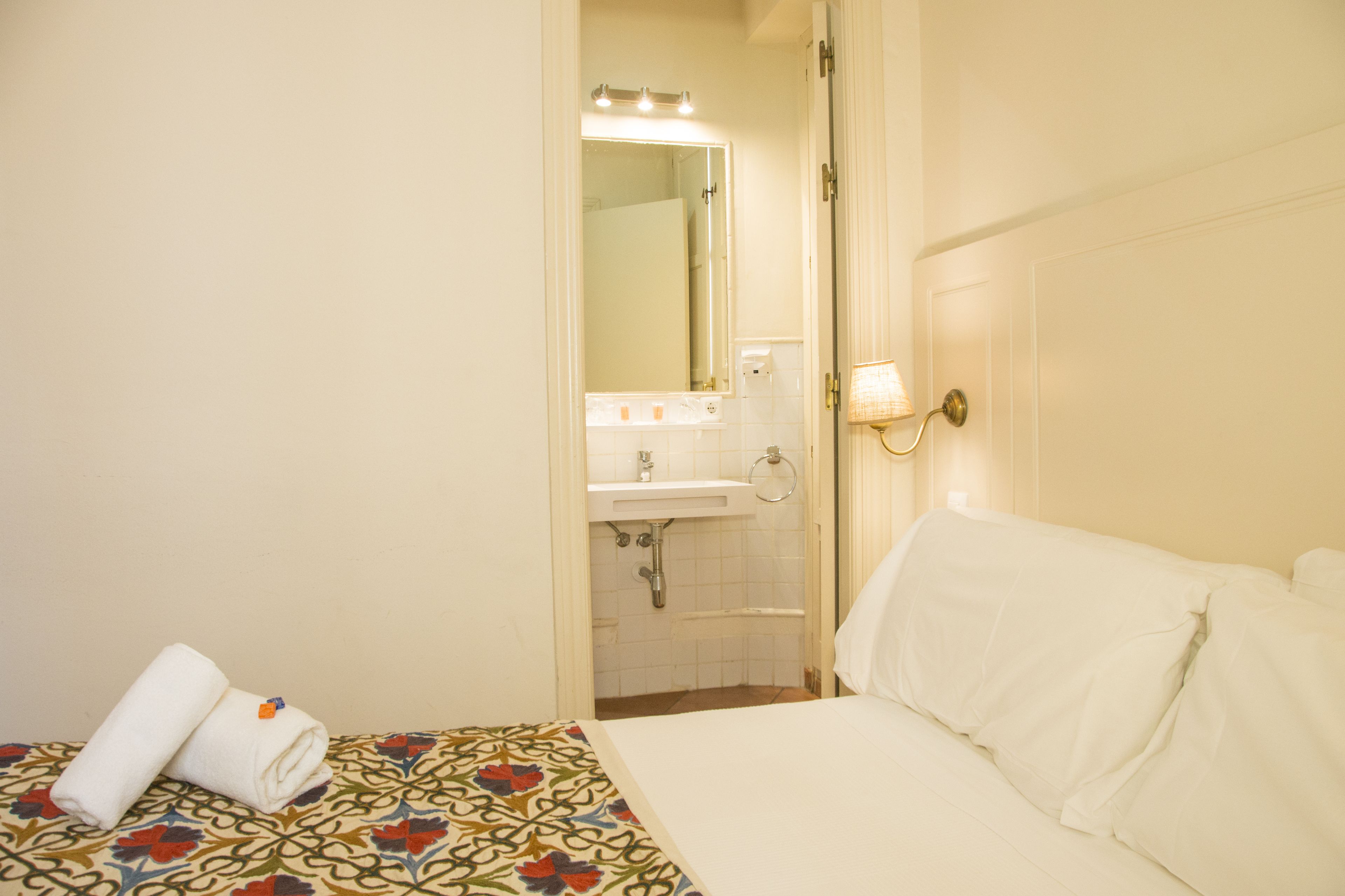 Habitación de hotel con una cama, una toalla doblada con cuidado, un baño con lavamanos y espejo, y una lámpara de pared.