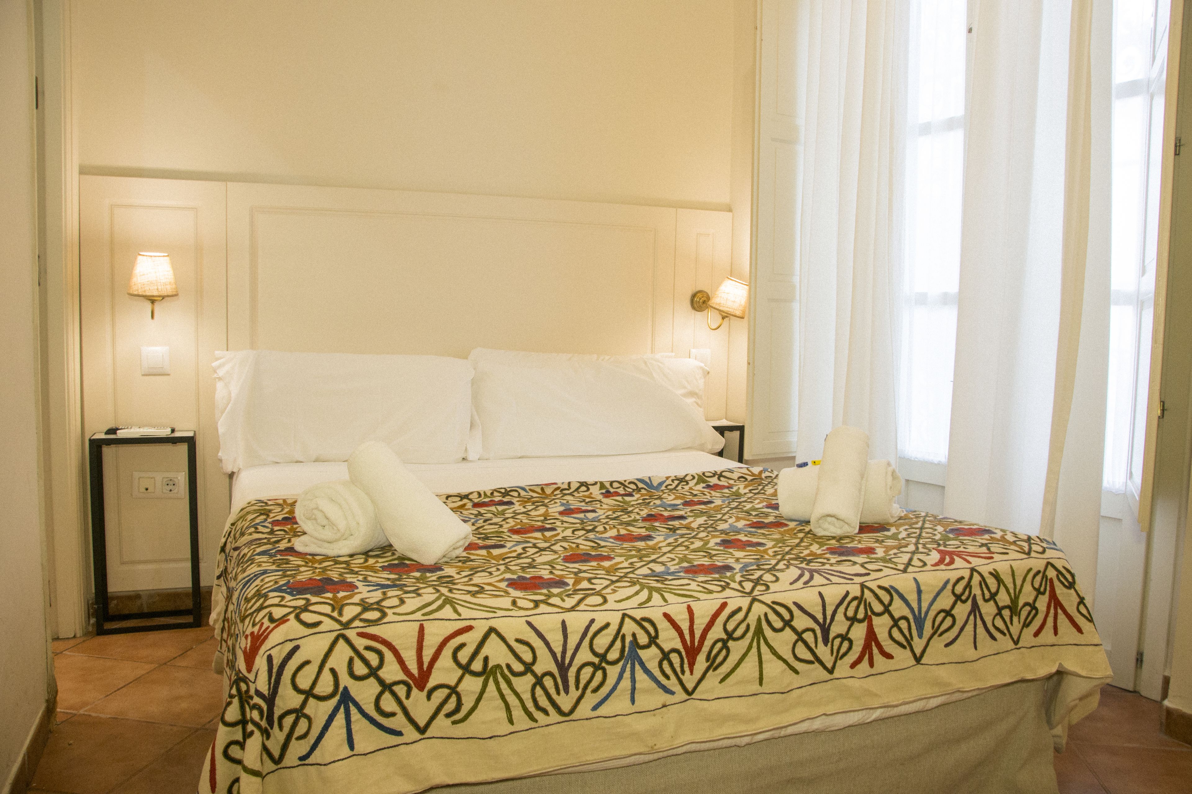 Una cama bien hecha con una colcha estampada, dos lámparas de noche y una ventana con cortinas de gasa.