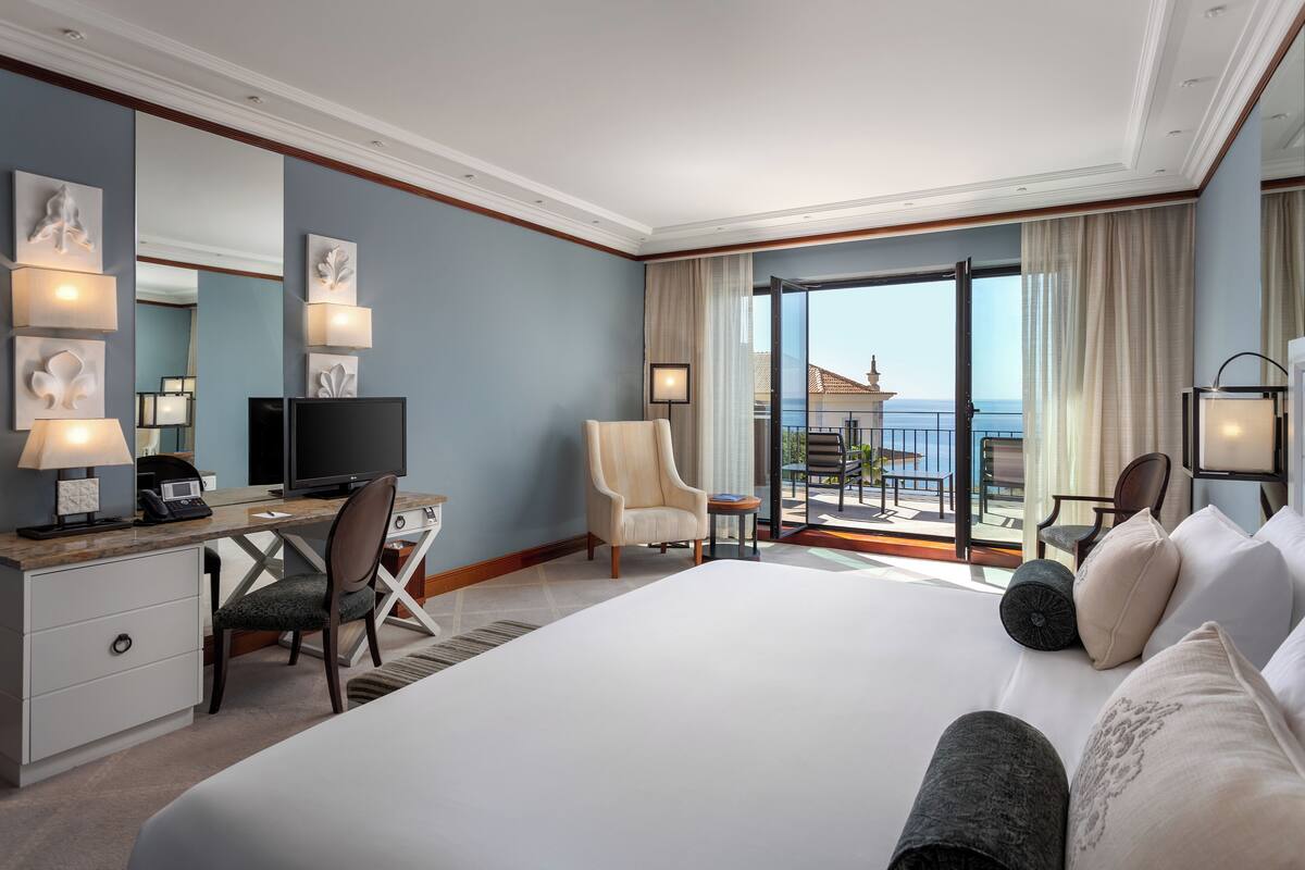 Ein Hotelzimmer mit einem großen Bett, einem Schreibtisch mit Computer, einem Sessel, einem Fernseher und einem Balkon mit Meerblick.