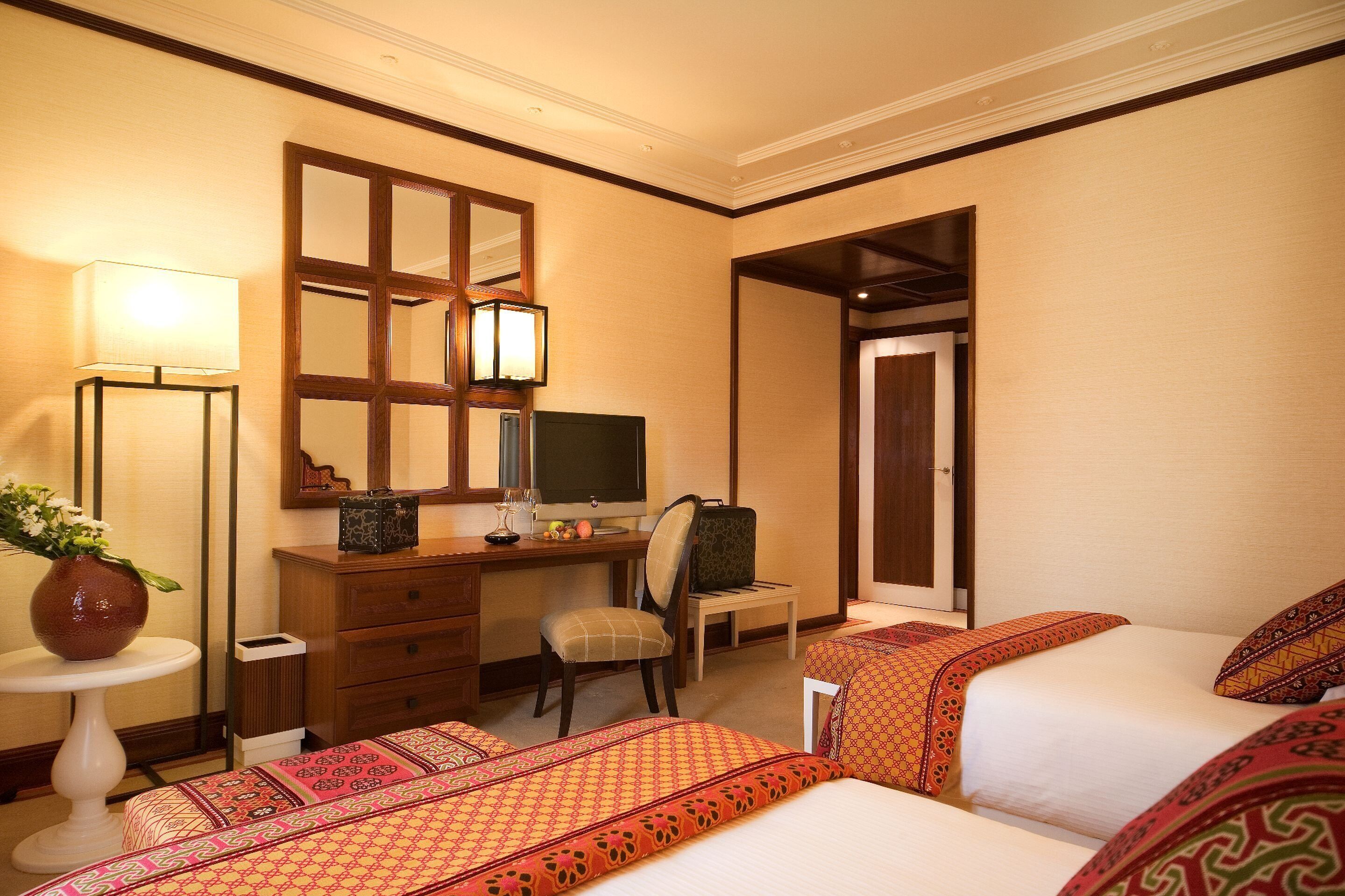 Quarto de hotel com duas camas, uma escrivaninha com computador, uma cadeira, um abajur e um espelho.