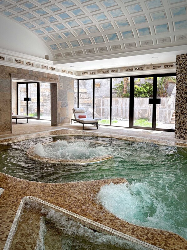 A spa area with a hot tub, a lounge chair, and a view of the outdoors through large windows. - Grande Real Villa Itália Hotel & Spa - A Leading Hotel of the World (Cascais)