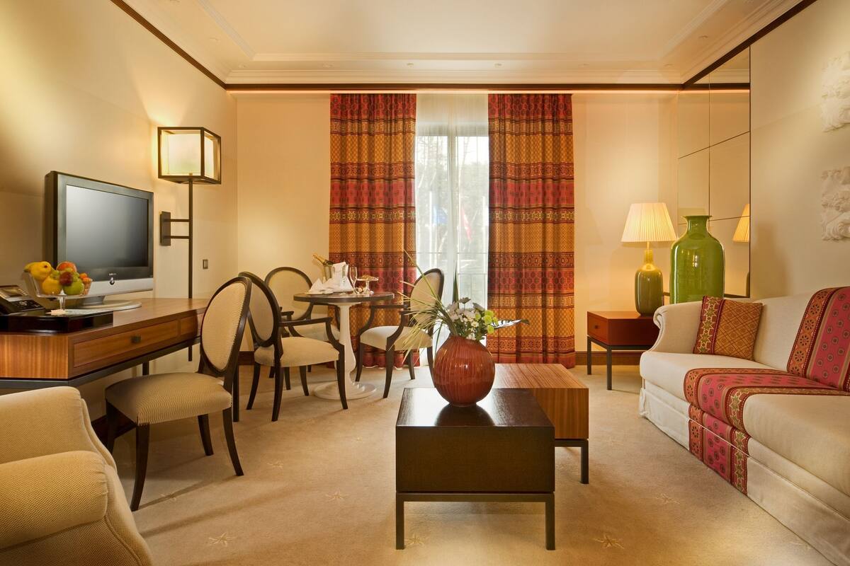 Ein Hotelzimmer mit Fernseher, einem Schreibtisch mit Blumenvase, einer Sitzecke und einem Essbereich mit Stühlen und Tisch.