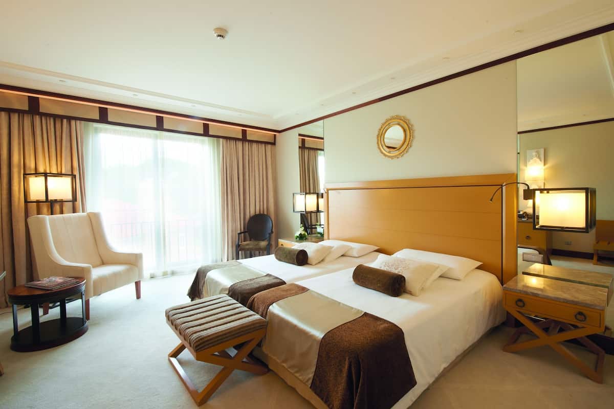Ein Hotelzimmer mit zwei Betten, einer Sitzecke mit Sessel und kleinem Tisch, einem großen Fenster mit Vorhängen und einem Spiegel an der Wand.