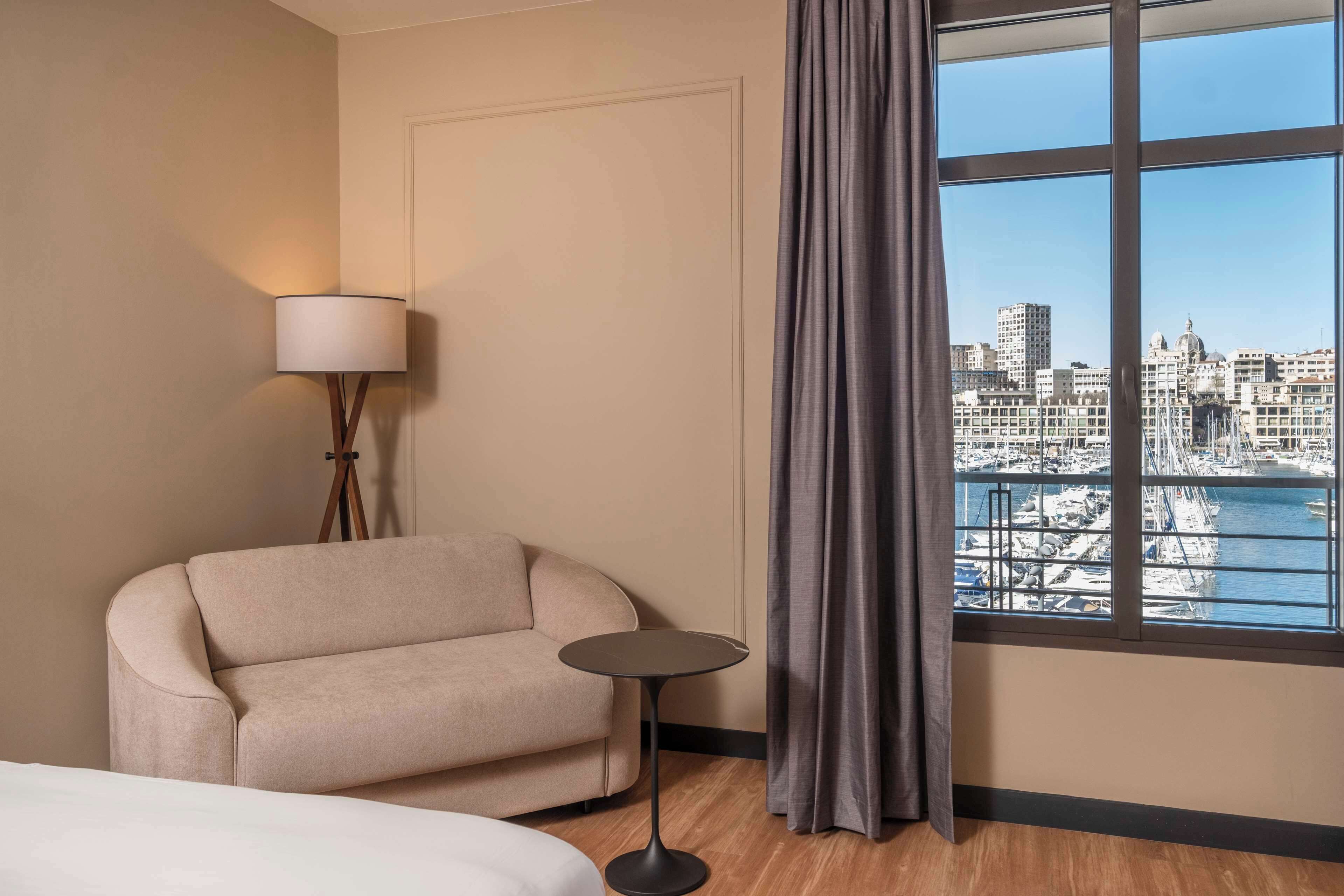 Habitación de hotel con sofá, una mesita redonda, lámpara y un ventanal con vistas a la ciudad.
