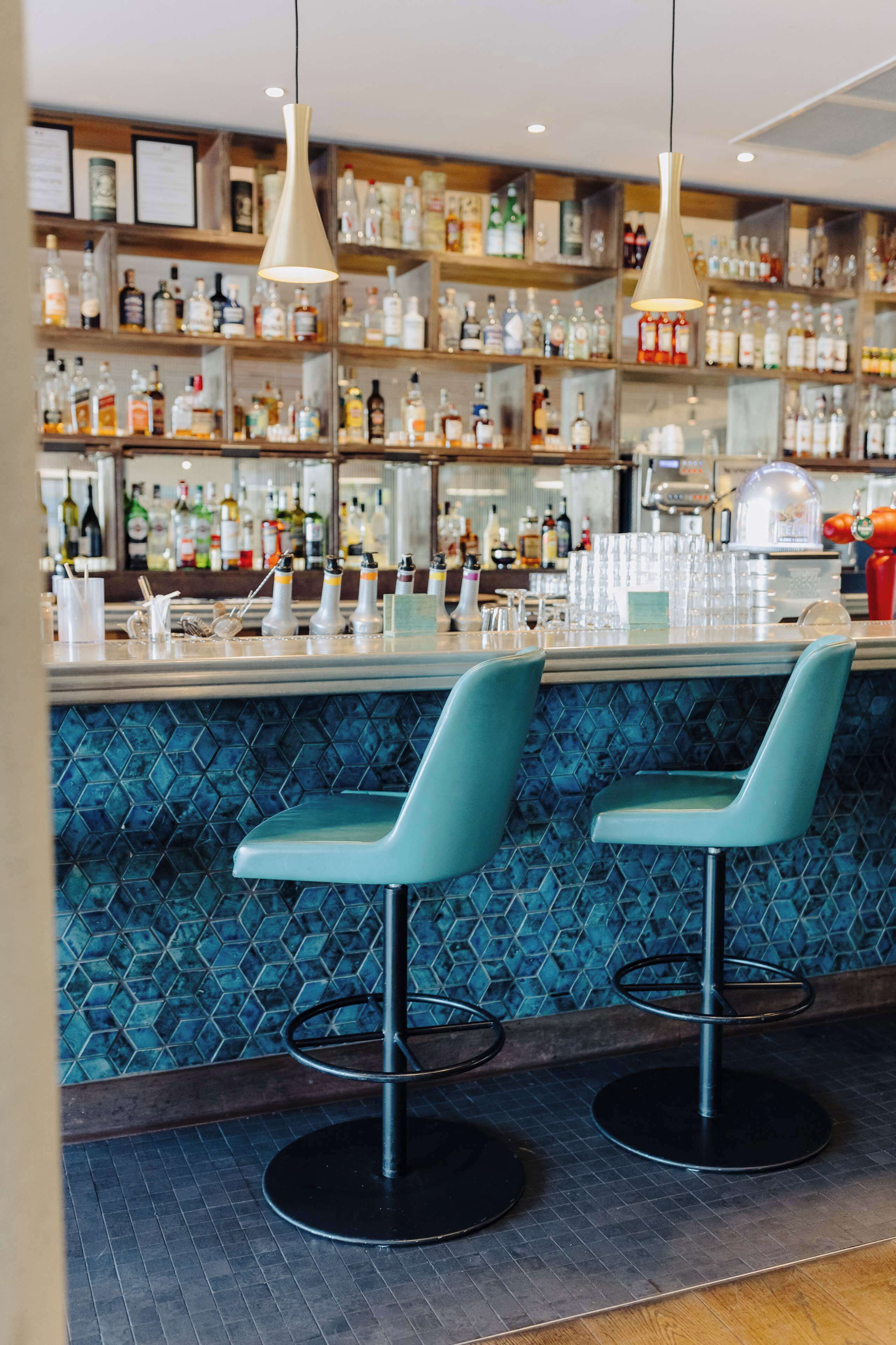 Un espace bar avec deux tabourets, un comptoir avec des verres et des bouteilles, et des étagères remplies de boissons alcoolisées.