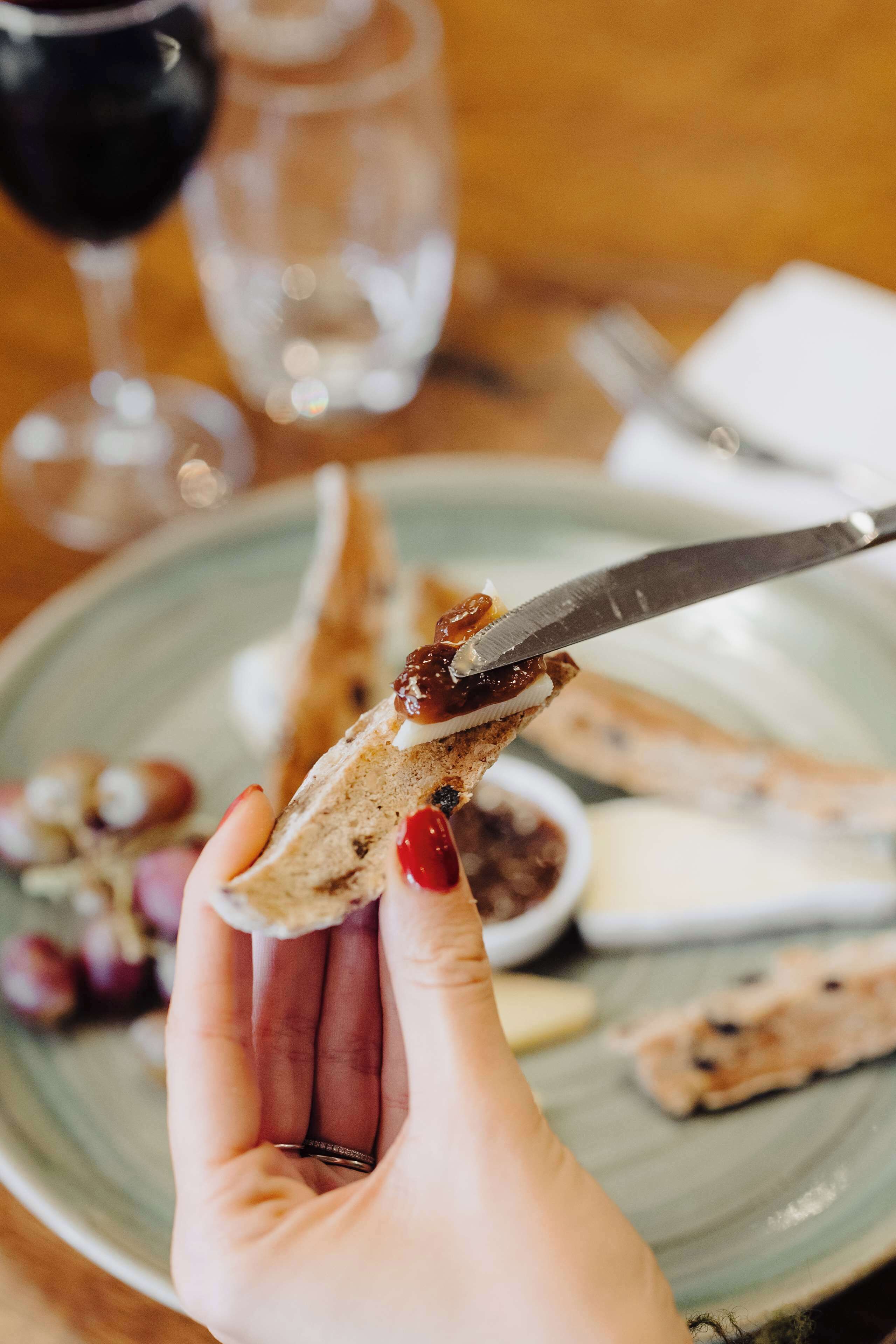 A hand holding a piece of bread with a spread, a knife, and a plate with other food items.