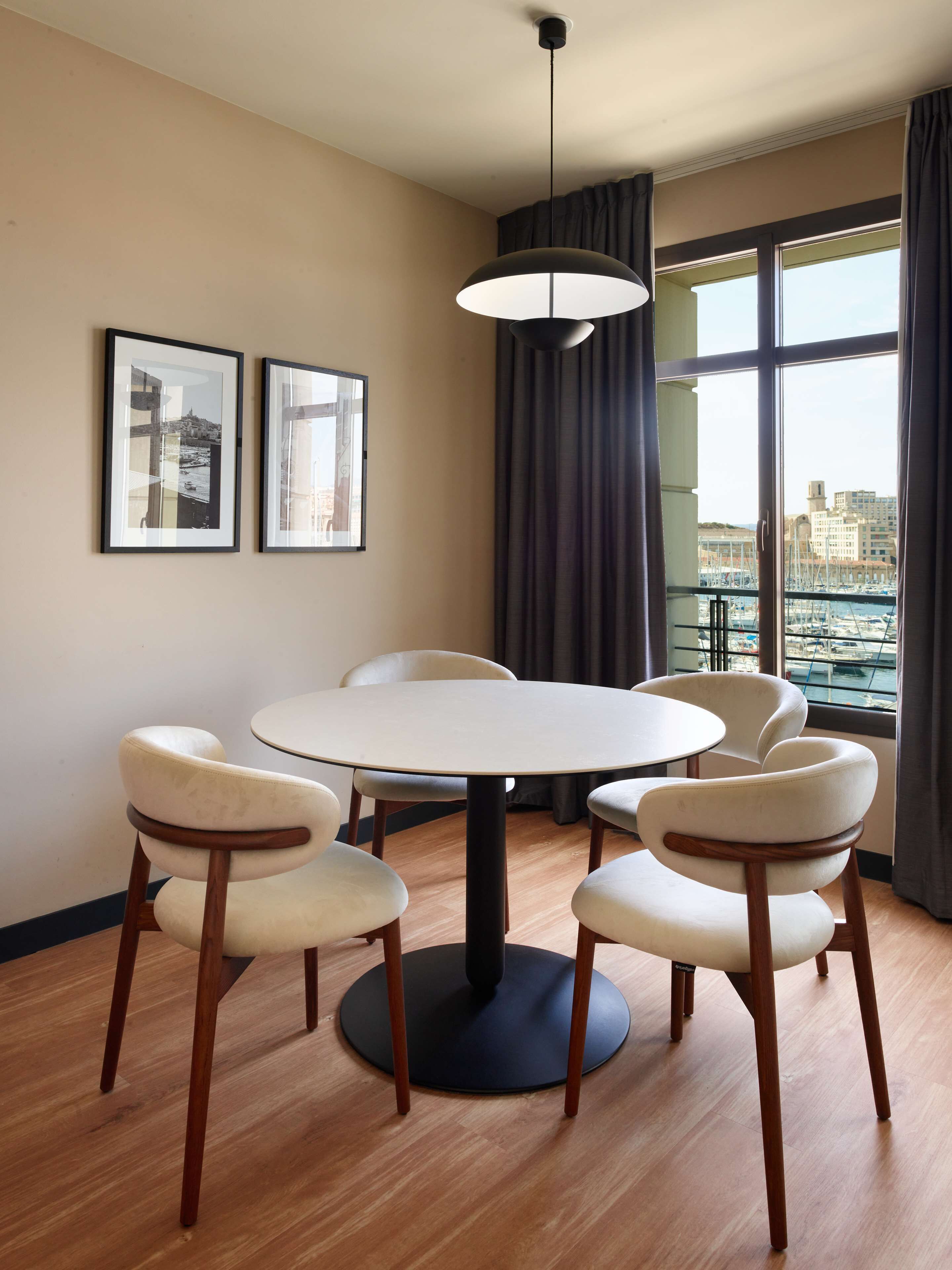 A modern dining area with a round table and six chairs, framed pictures on the wall, a large window with a view of the city, and a hanging light fixture.