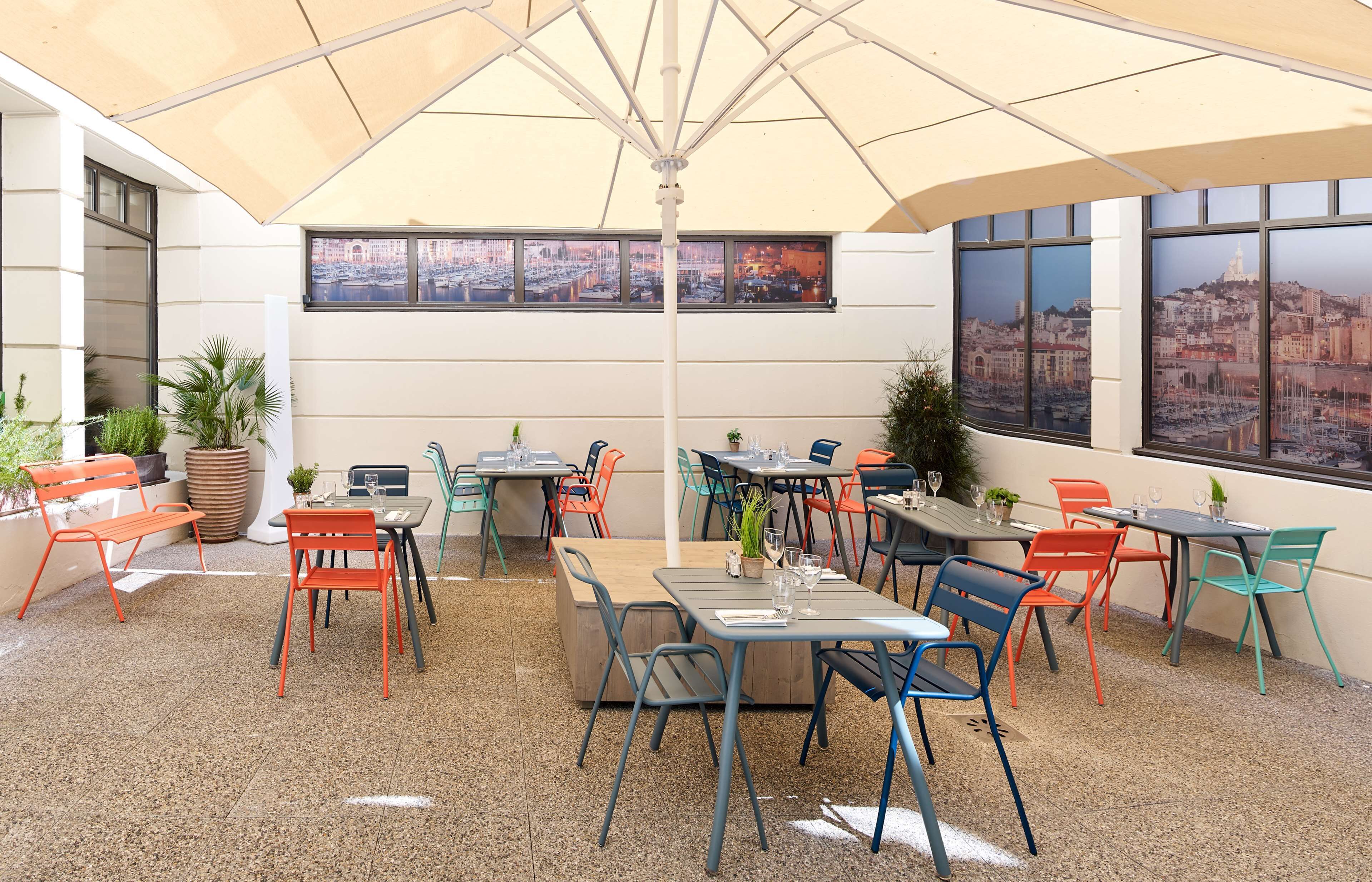 Un café en plein air avec de nombreuses tables et chaises, un grand parasol et une vue sur la ville à travers les fenêtres.