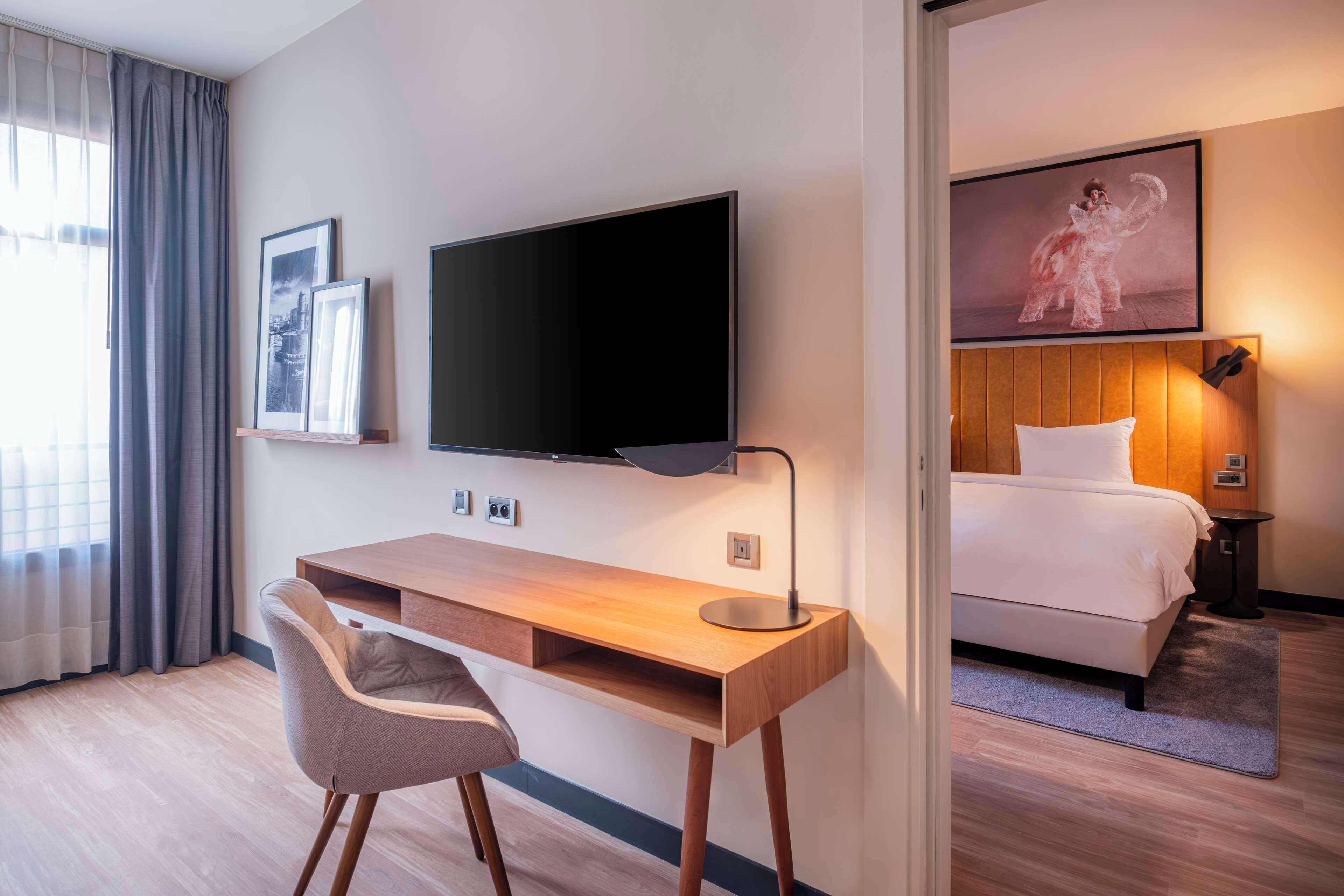 Ein Hotelzimmer mit Holzschreibtisch, Flachbildfernseher, Bett mit weißer Bettwäsche und einem gerahmten Bild an der Wand.