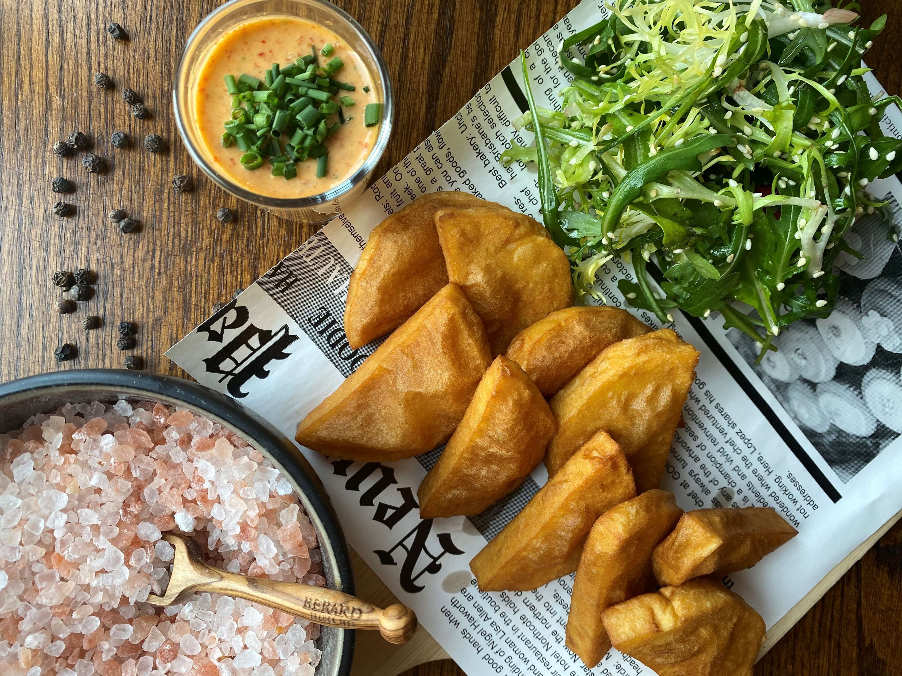 A plate of golden-brown fried items, a bowl of green onion dip, a pile of green salad, and a bowl of pink salt crystals on a wooden table with a newspaper underneath.