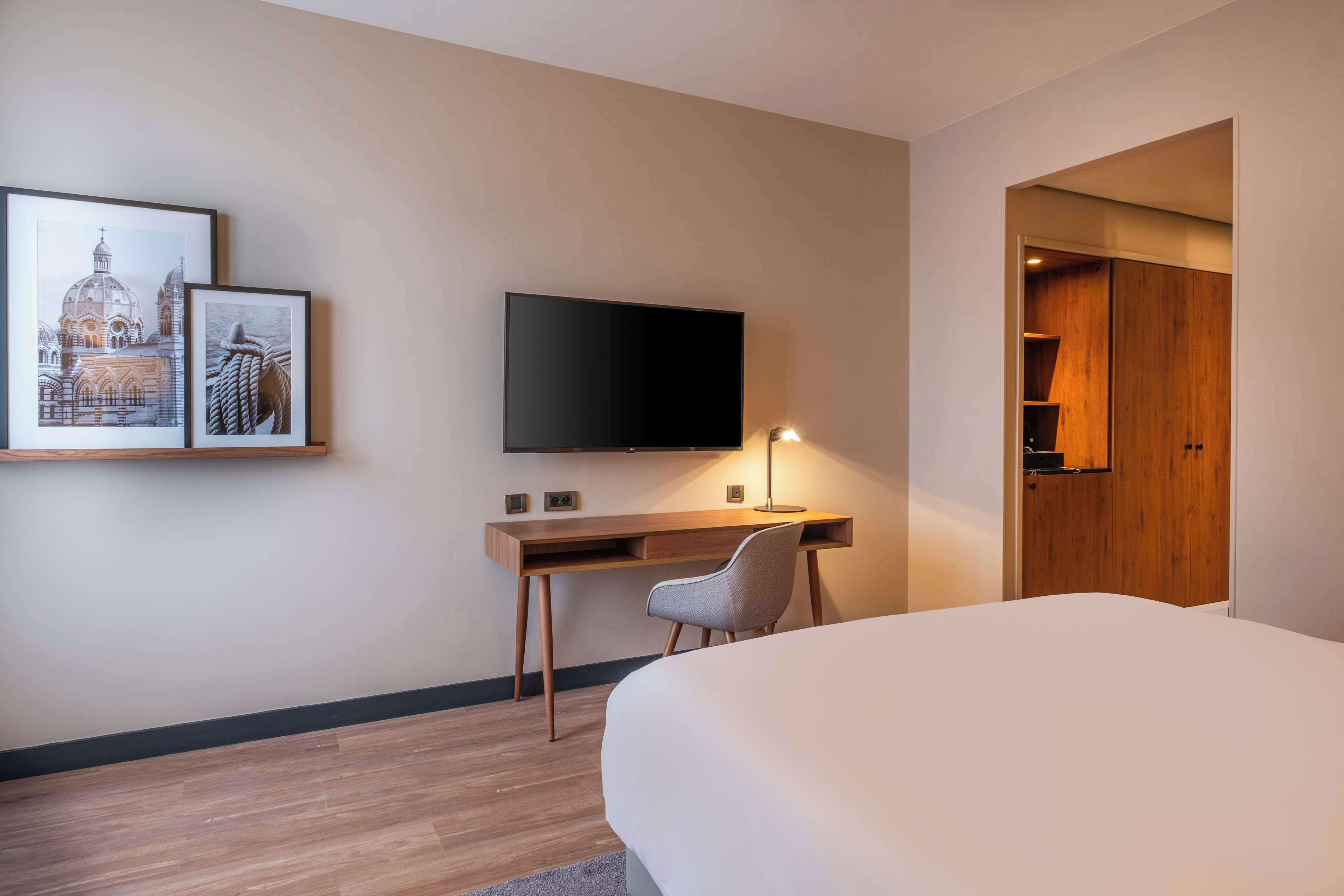A hotel room with a bed, a desk with a chair, a flat-screen TV, and two framed pictures on the wall.
