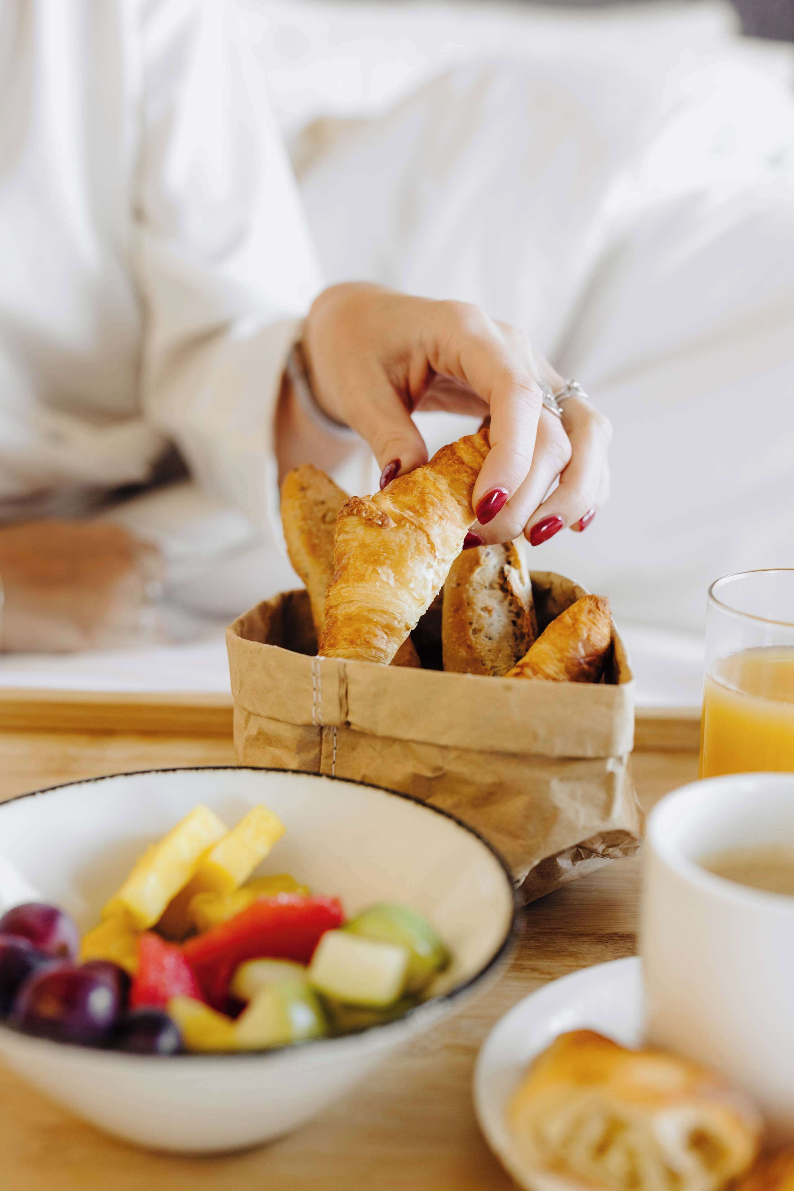 A person holding a croissant from a paper bag, a bowl of fruit, and a glass of orange juice on a table.
