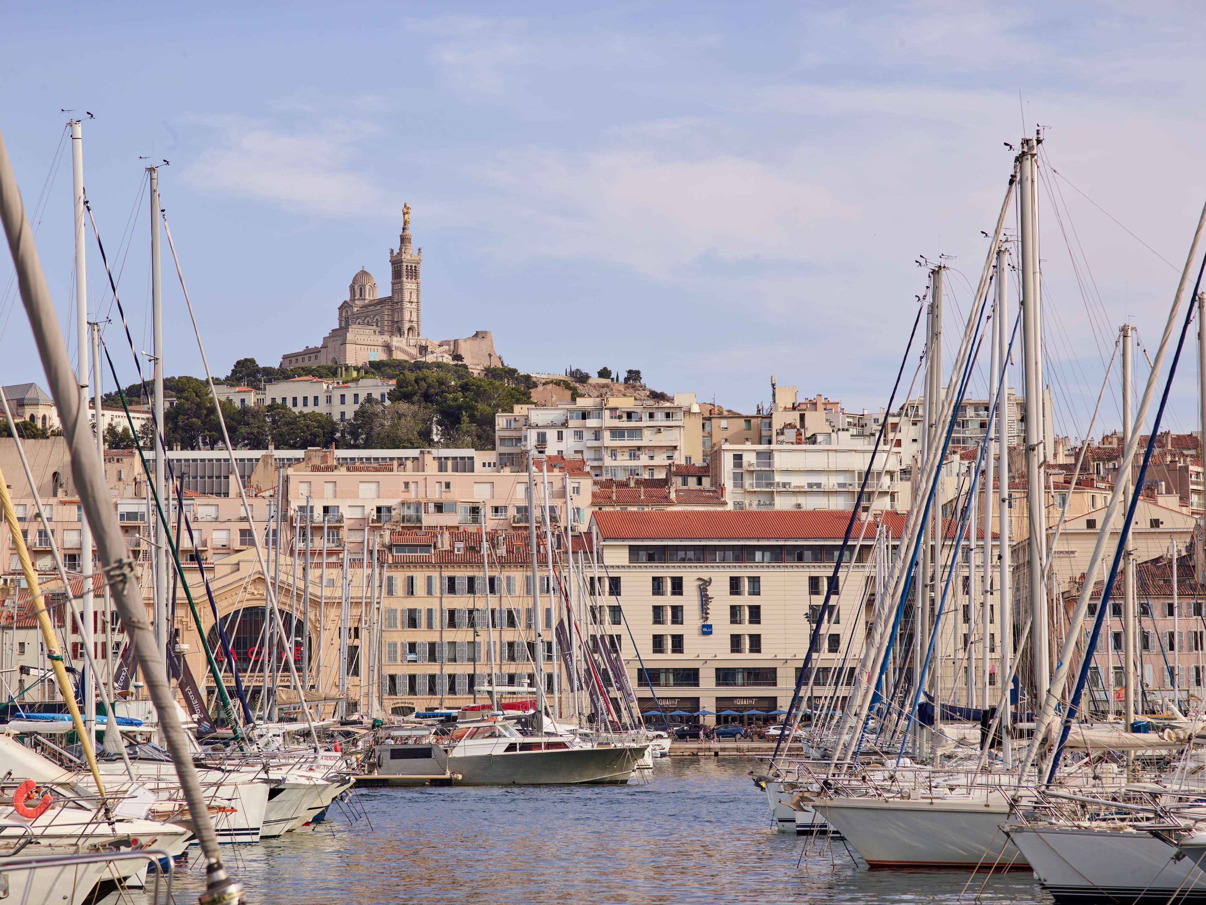 Un port de plaisance où sont amarrés de nombreux bateaux, un bâtiment imposant surmonté d’un dôme et d’une statue en arrière-plan, et un paysage vallonné.