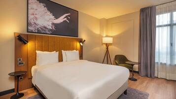 Habitación de hotel con una cama grande, cabecera de madera, lámpara de pared, una mesita, una silla y una ventana con cortinas.