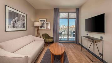 Quarto de hotel moderno com sofá, uma mesa redonda pequena, televisão e uma janela grande com vista para a cidade.