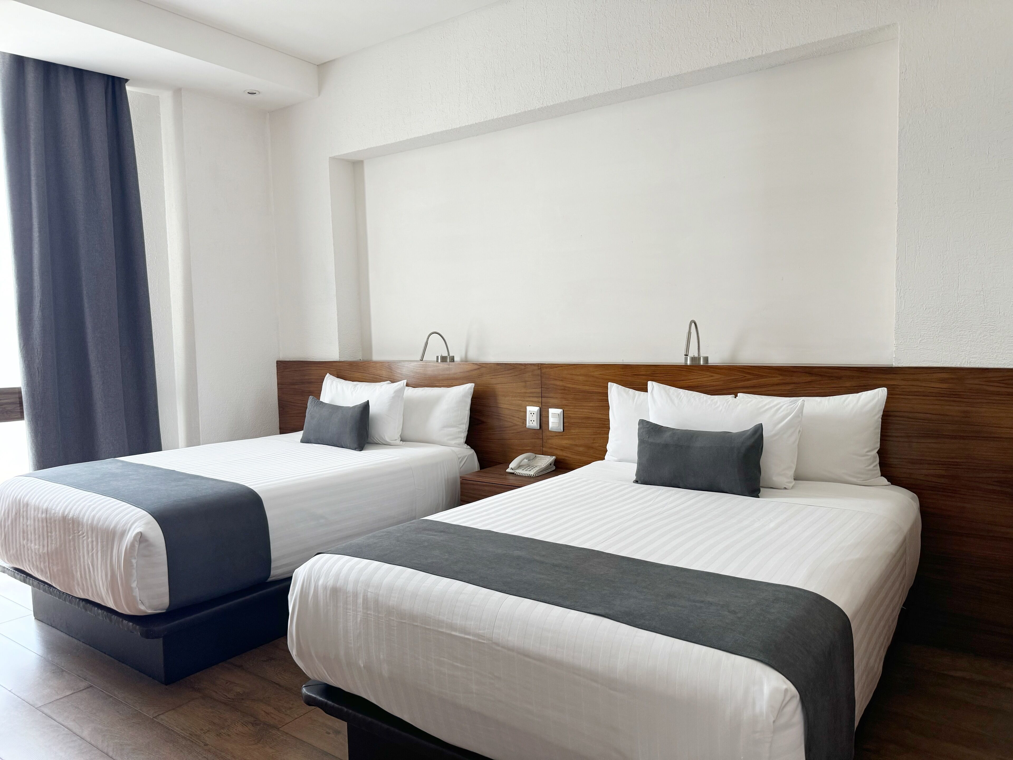 Une chambre d’hôtel avec deux lits, une tête de lit en bois, une table de chevet avec un téléphone et une fenêtre avec des rideaux.