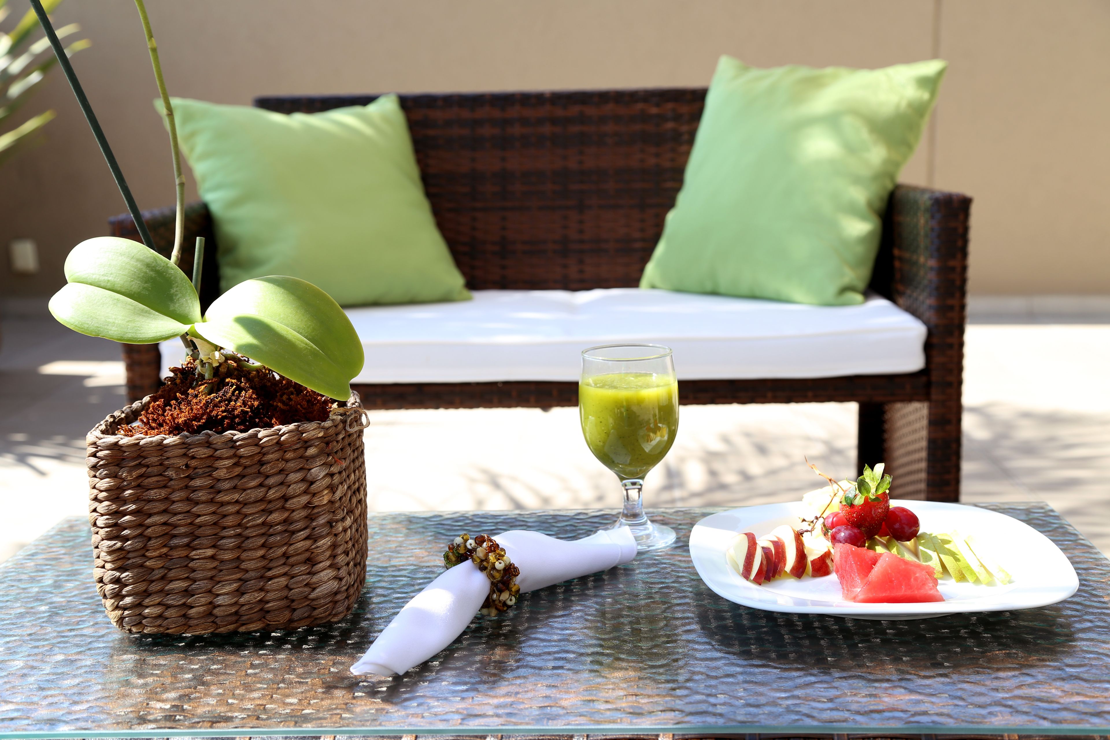 Een rieten pot met een plant, een glas groene drank, en een bord met fruit en vlees op een glazen tafel.