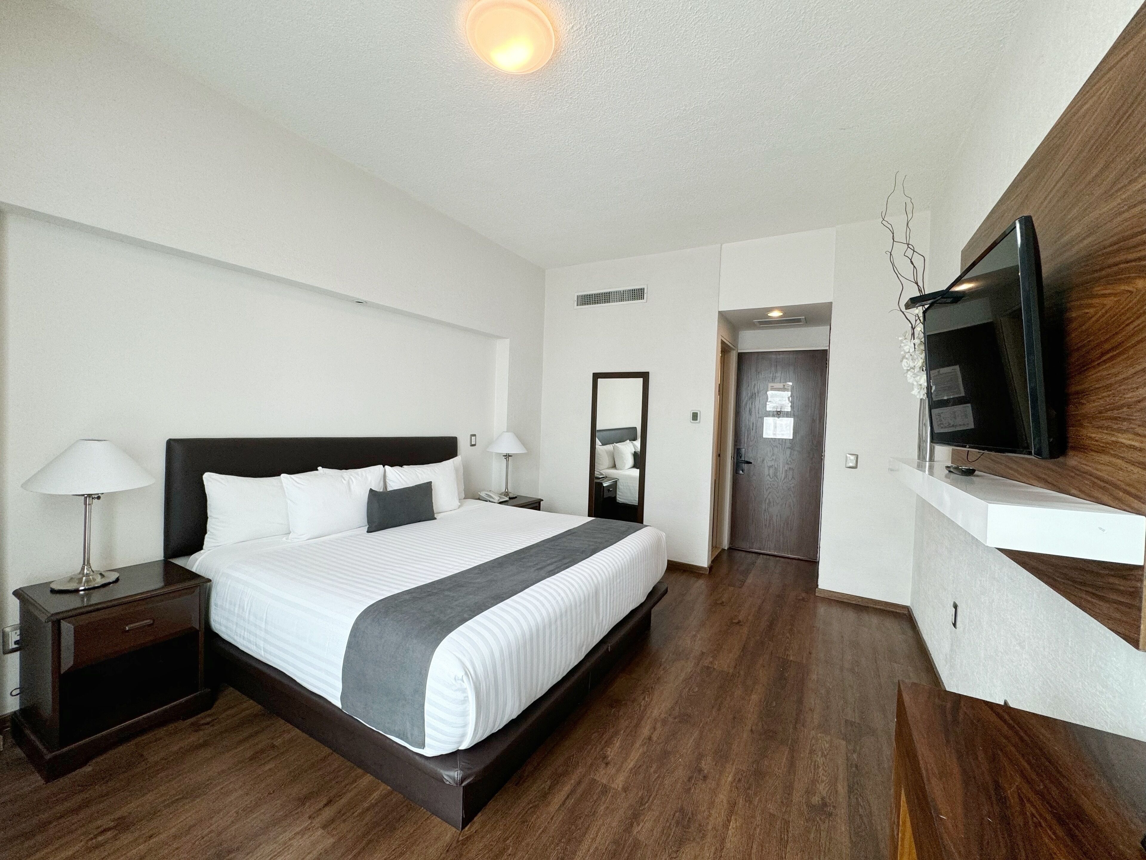 A modern hotel room with a large bed, bedside tables, a flat-screen TV mounted on the wall, and a wooden floor.