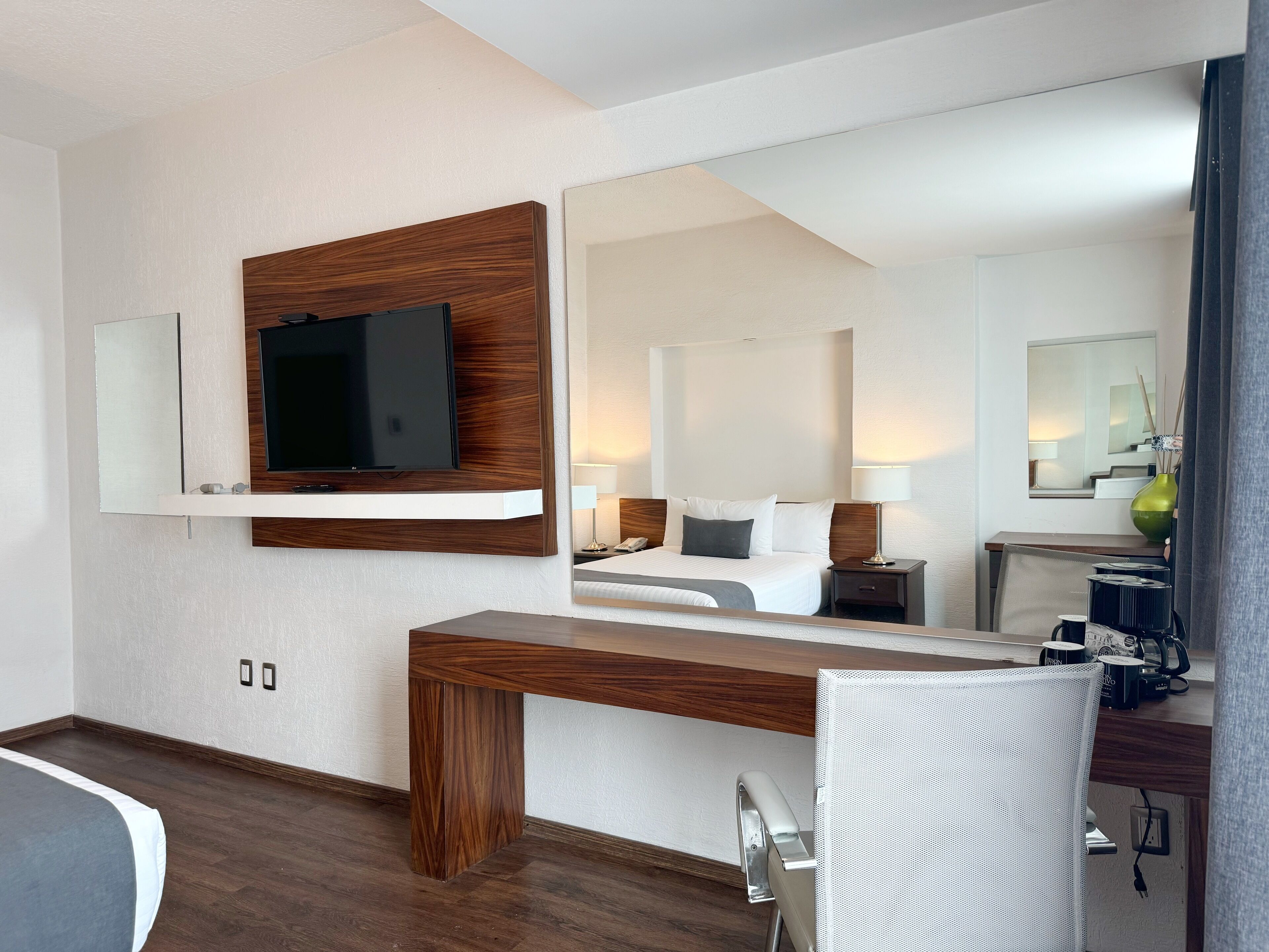 Una camera d'albergo moderna con una scrivania in legno, una TV a schermo piatto fissata al muro, uno specchio grande e un letto confortevole con un comodino.