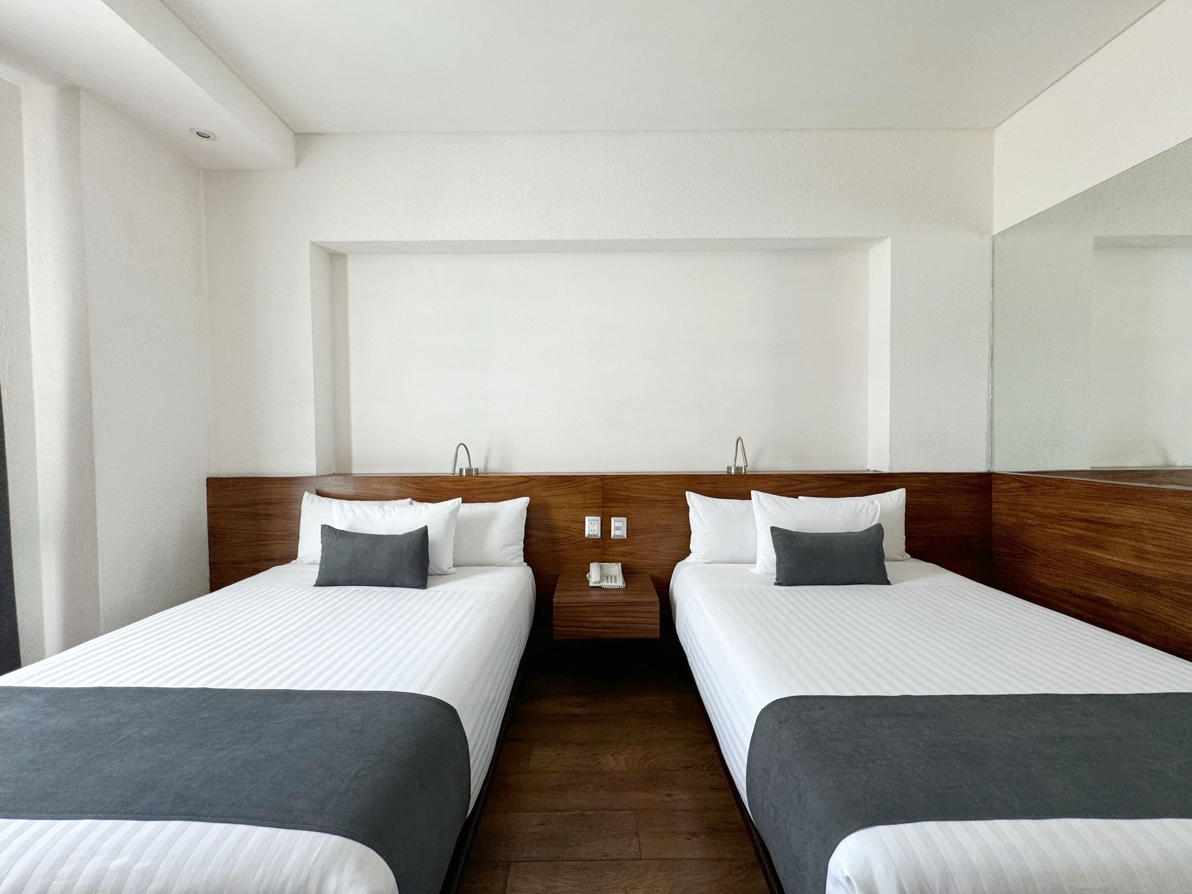 Une chambre d’hôtel avec deux lits simples, des têtes de lit en bois, du linge de lit blanc et une table de chevet avec un téléphone et une lampe.