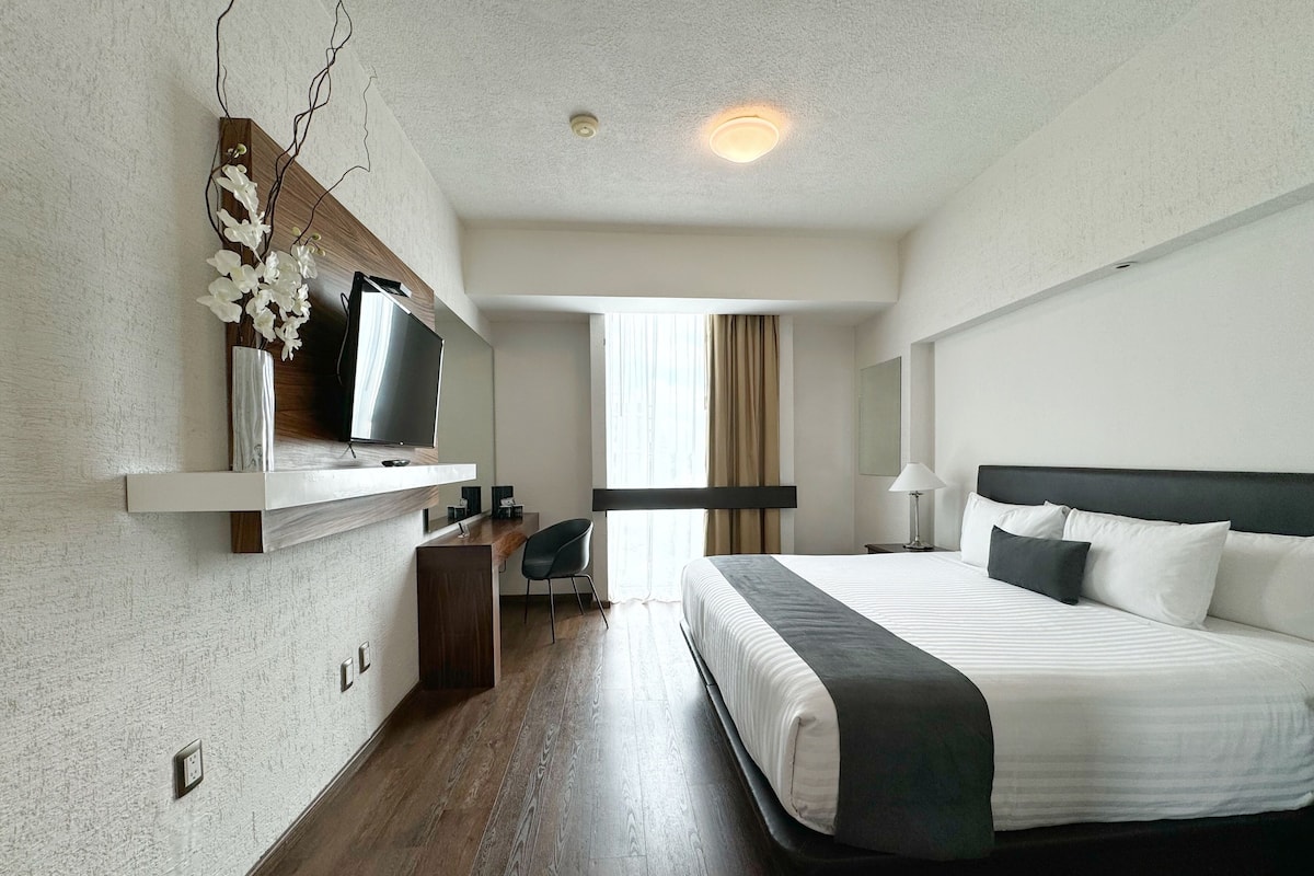Una habitación de hotel moderna con una cama grande, un escritorio con una silla, televisión y una ventana con cortinas.