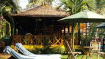 A covered outdoor seating area with a thatched roof, wooden furniture, and a palm tree in the background.