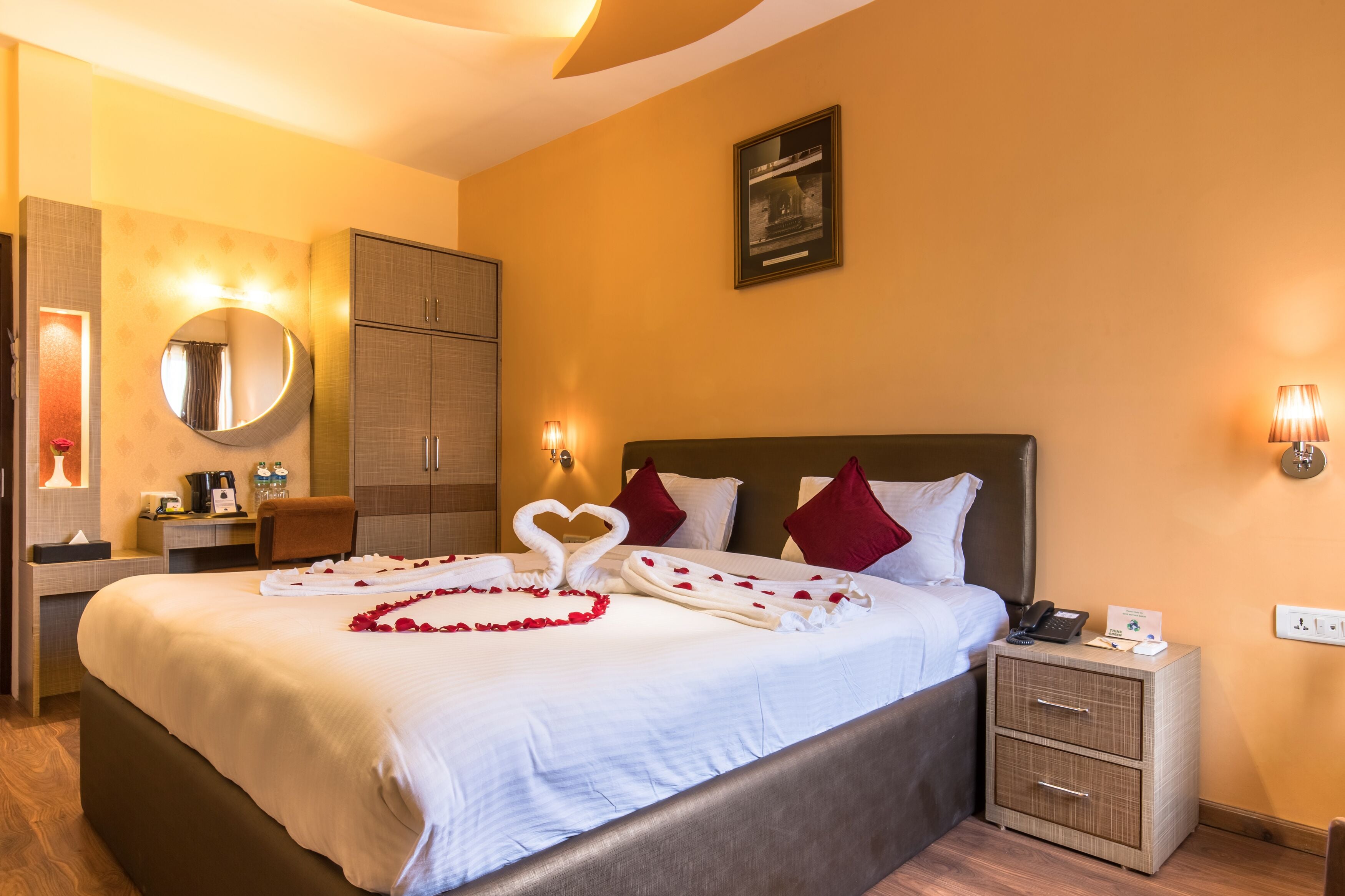 A hotel room with a large bed, a bedside table, a nightstand, a mirror, a wardrobe, and a framed picture on the wall.
