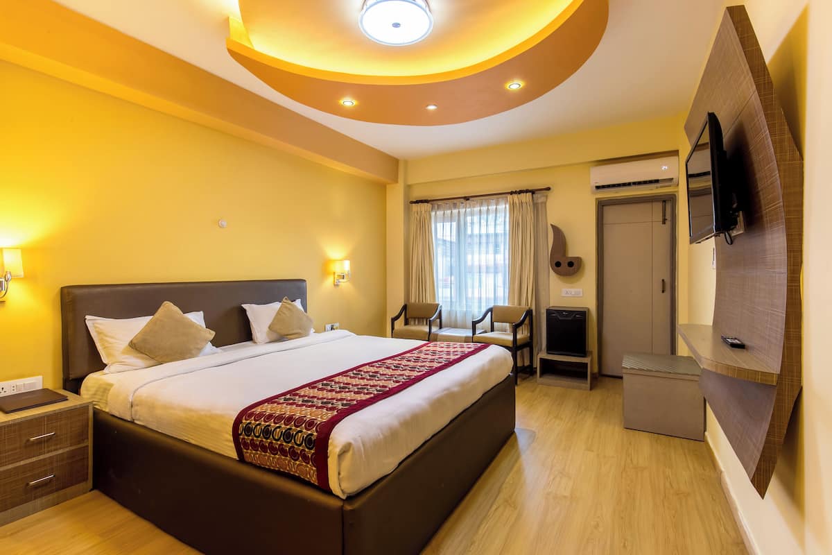 A hotel room with a large bed, bedside lamps, a flat-screen TV, a desk with a chair, and an air conditioning unit.